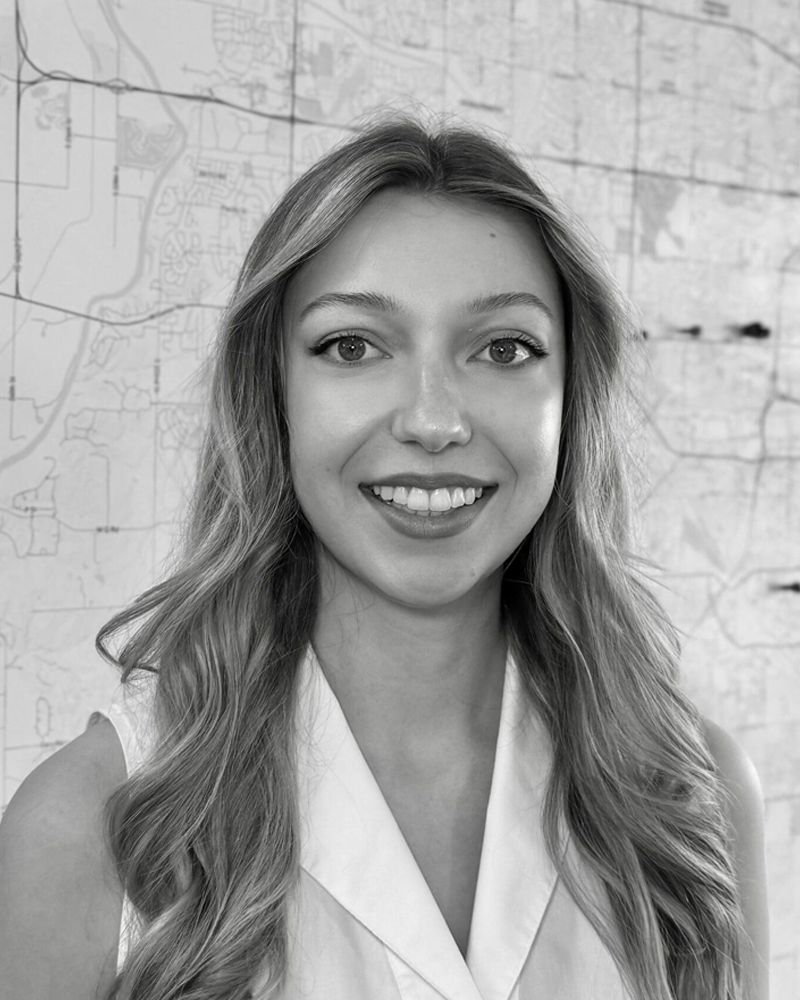 Black and white portrait of a young woman with long wavy hair, smiling, standing in front of a map.