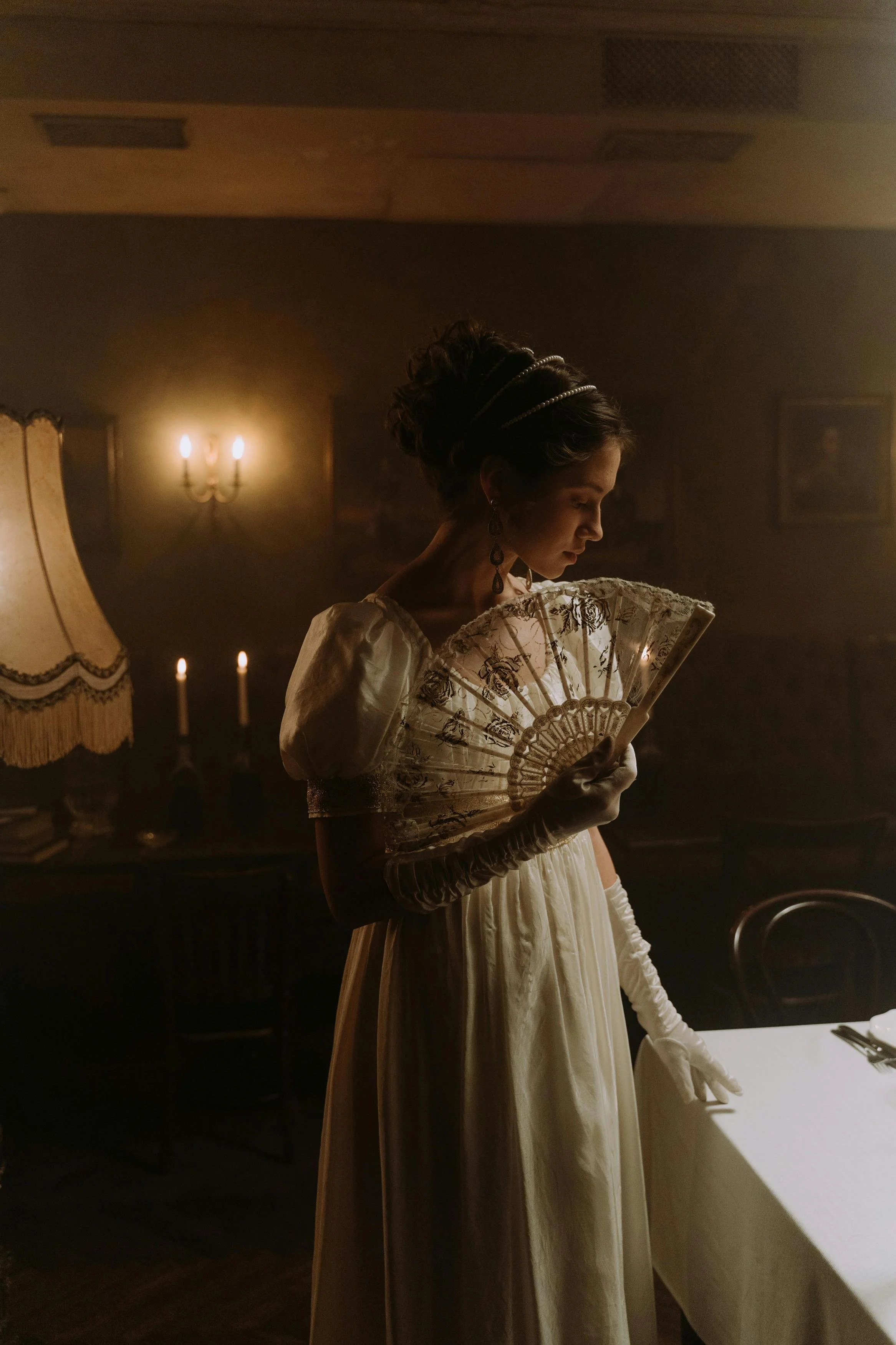 A woman dressed in a vintage cream-colored gown with puffed sleeves, wearing long gloves and ornate earrings, holds a decorative lace fan and stands in a dimly lit, antique-styled room. She appears to be looking down at the fan.