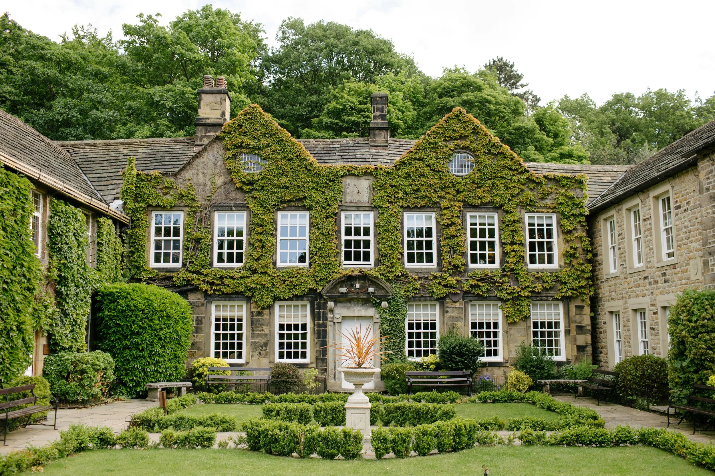 A historic stone mansion covered in green ivy, with numerous white-framed windows and a landscaped garden in the foreground, including benches, bushes, a small lawn, and a central stone fountain.