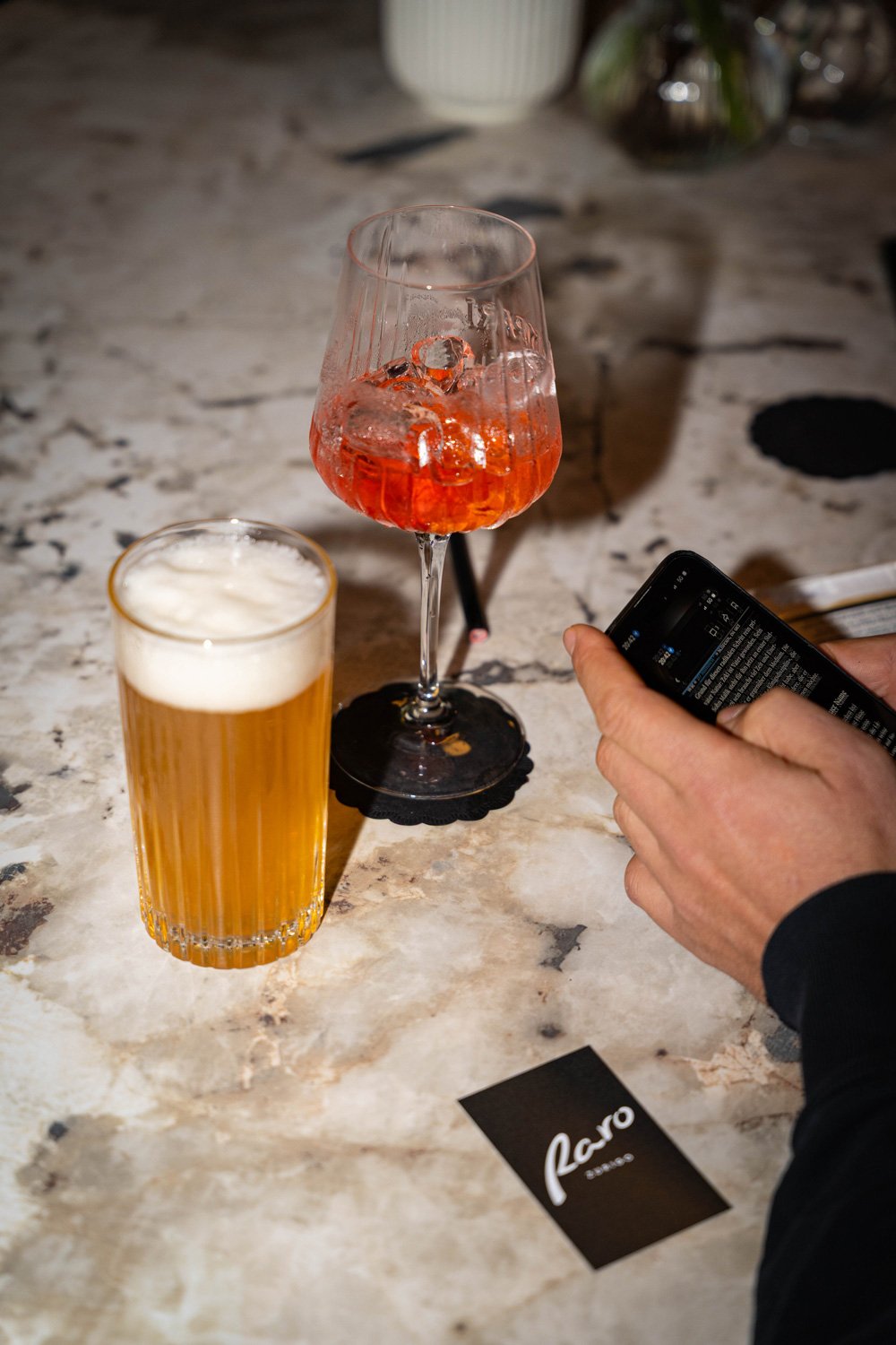 Zwei Getränke, ein Bier und ein Cocktail auf einem Marmor-Tisch, eine Person benutzt ein Smartphone, im Hintergrund eine Pflanze.