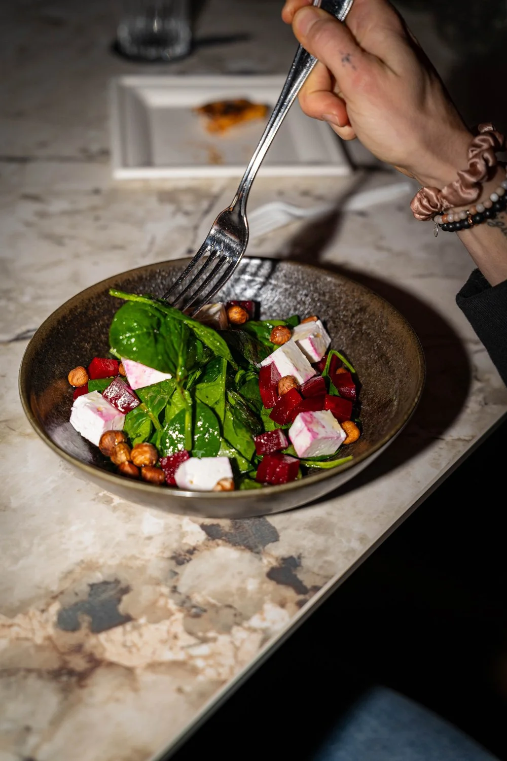 Schüssel mit Salat, bestehend aus Blattspinat, gewürfeltem Ziegenkäse, rotem Rüben, Haselnüssen, auf einem Marmortisch. Jemand hält eine Gabel über die Salatschüssel.