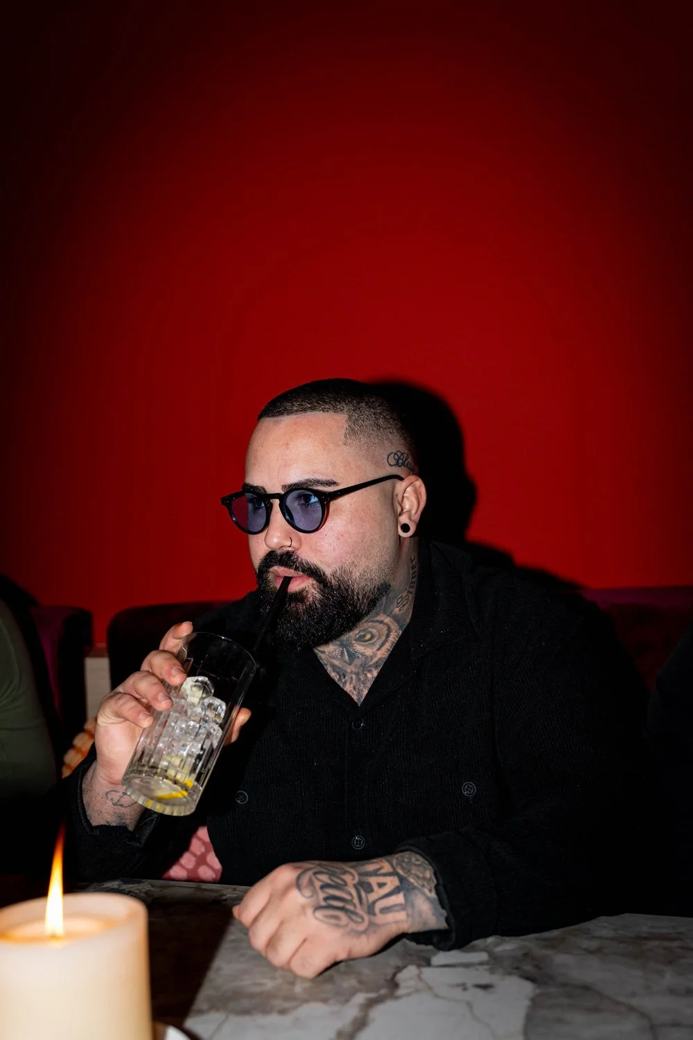 Ein Mann mit Tattoos, dunklem Bart und schwarzen Haaren trinkt einen Drink mit einem schwarzen Strohhalm in einem Glas, trägt schwarze Brillen und sitzt an einem Tisch mit einer Kerze vor einem roten Hintergrund.