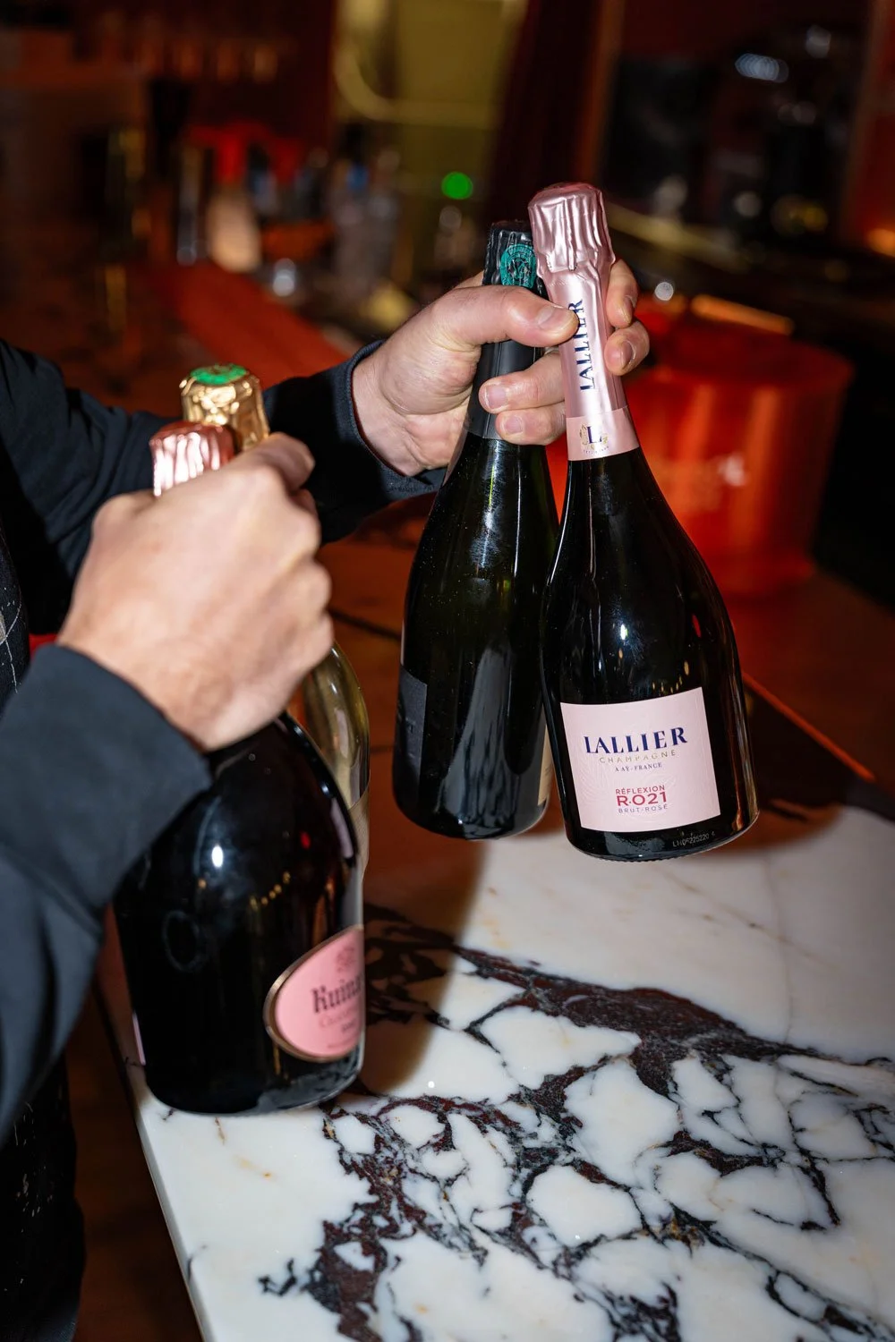 Person öffnet eine Flasche Champagner auf einem Marmortresen, weitere Flaschen im Hintergrund.