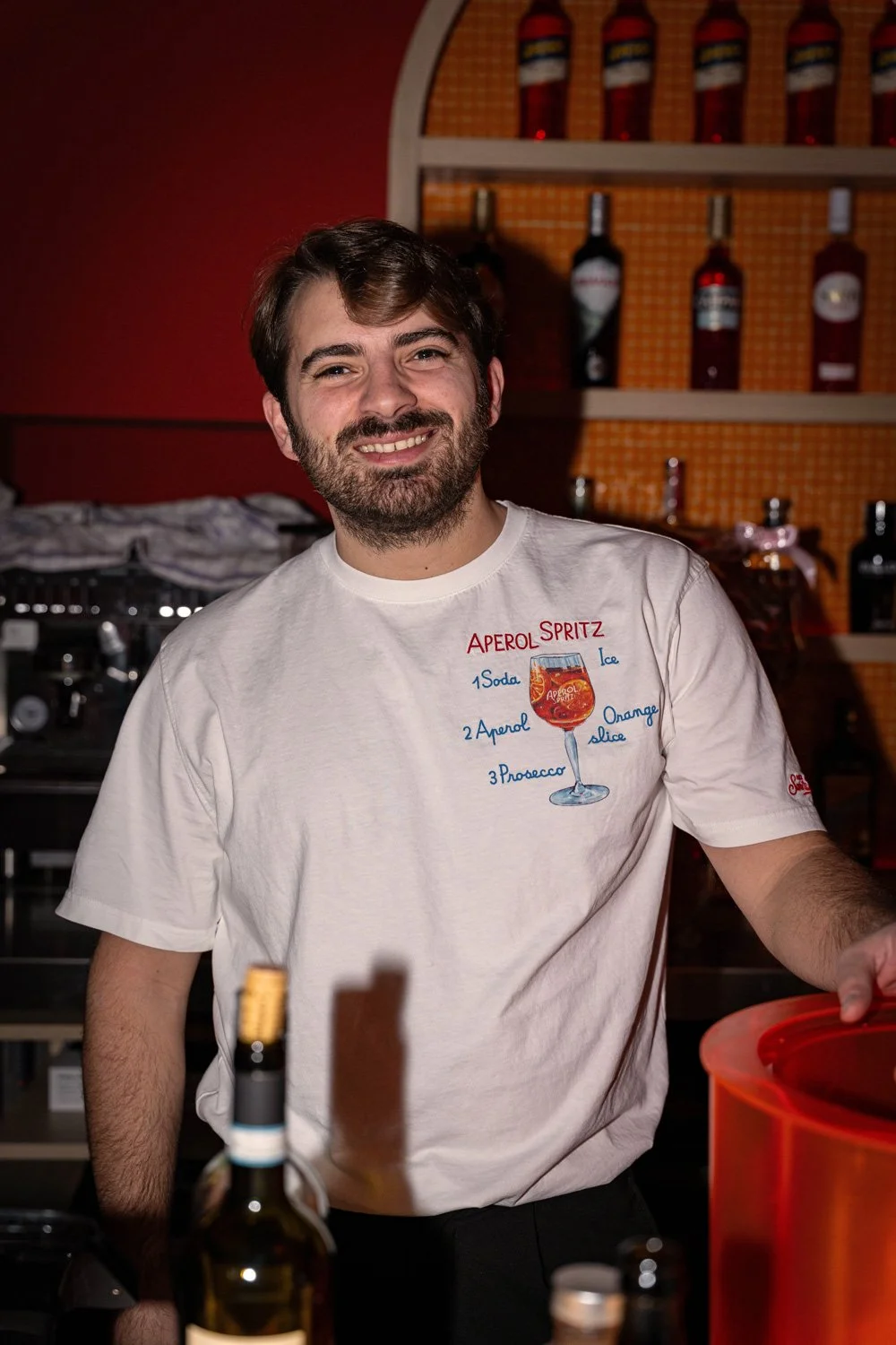 Ein lächelnder Mann mit Bart trägt ein weißes T-Shirt mit einem Rezept für Aperol Spritz, vor einer Bar mit Brennflaschen im Hintergrund.
