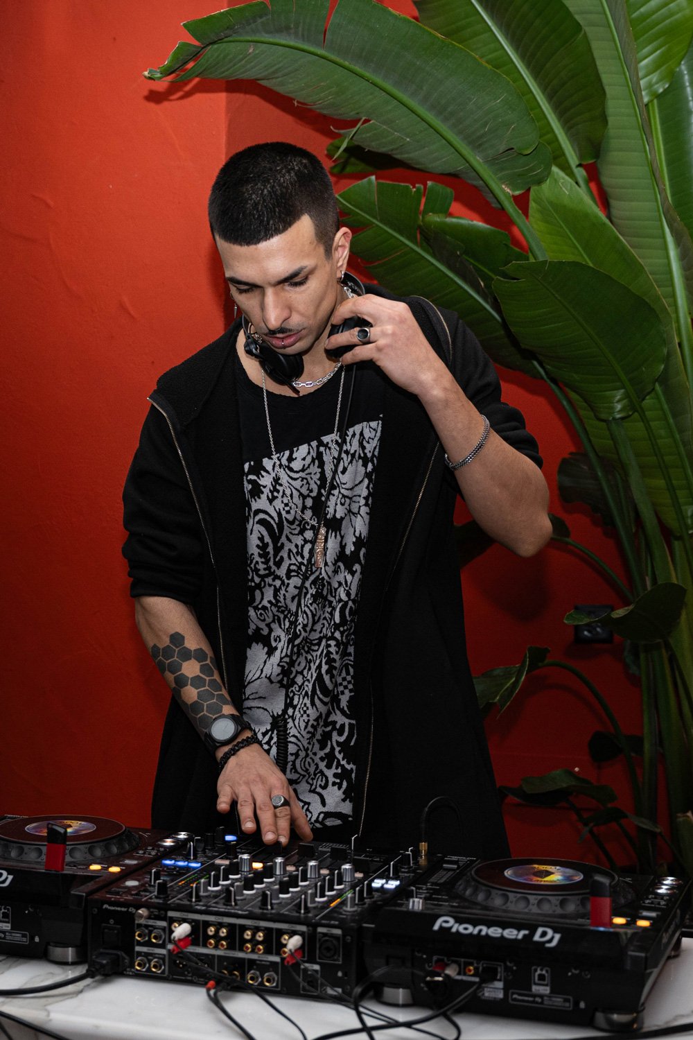 Ein junger Mann mit Short, Tattoos und Schmuck, der an einem DJ-Controller arbeitet und Kopfhörer trägt, vor einer roten Wand mit einer großen grünen Pflanze im Hintergrund.