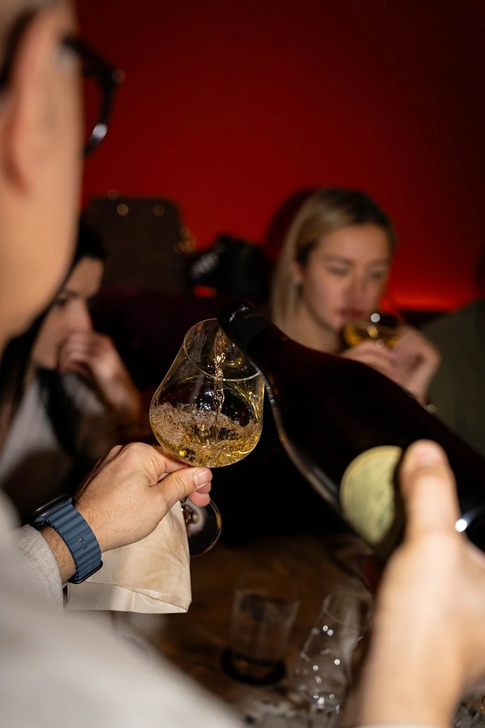 Menschen, die in einer Bar Champagner oder Sekt trinken, mit einer Hand, die ein Glas hält, im Vordergrund. Im Hintergrund ist eine Frau, die ebenfalls trinkt, die Szene hat ein dunkles, rotes Licht.