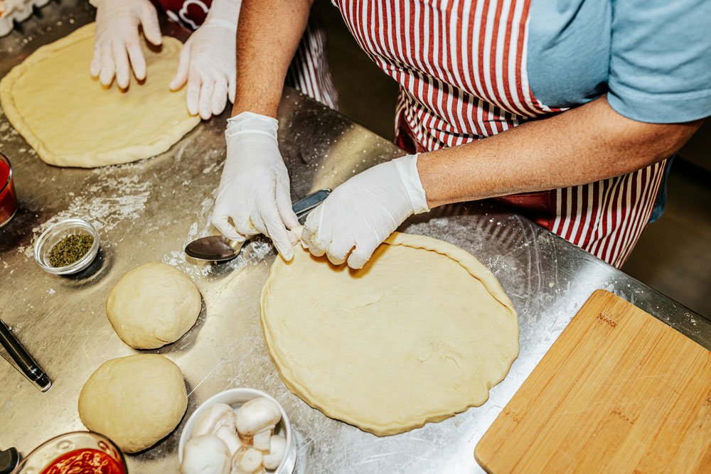 Pizzaiolo beim Teig ausrollen für die Zubereitung einer Pizza für ein italienisches Restaurant in Zürichc, mit weiteren Teigkugeln, Gewürzen und Küchenutensilien auf einem Metalltisch.