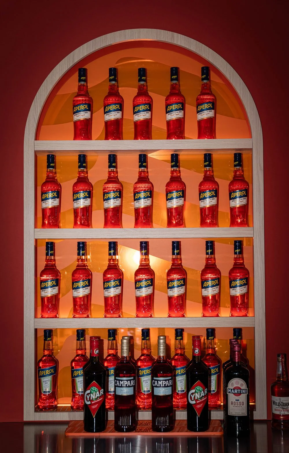 Regal mit verschiedenen alkoholischen Getränken, hauptsächlich Aperol-Flaschen, mit einem hinterleuchteten roten Hintergrund.