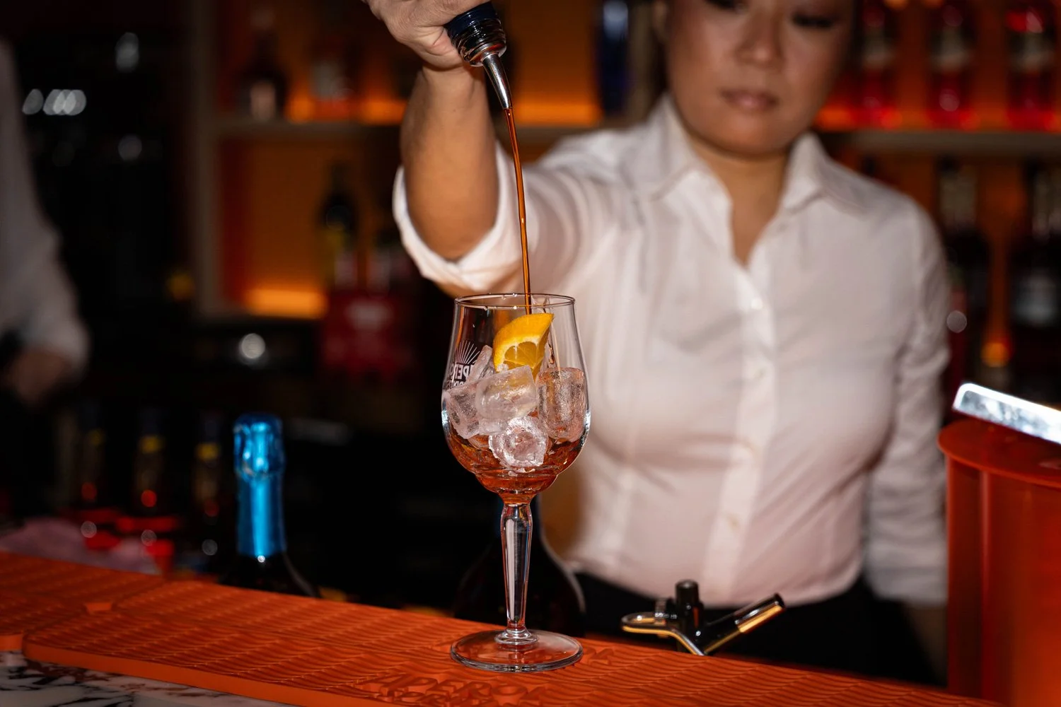 Mitarbeiterin beim Eingießen von Flüssigkeit in ein Weinglas mit Eiswürfeln, Zitrone und Bar-Umgebung im Hintergrund.