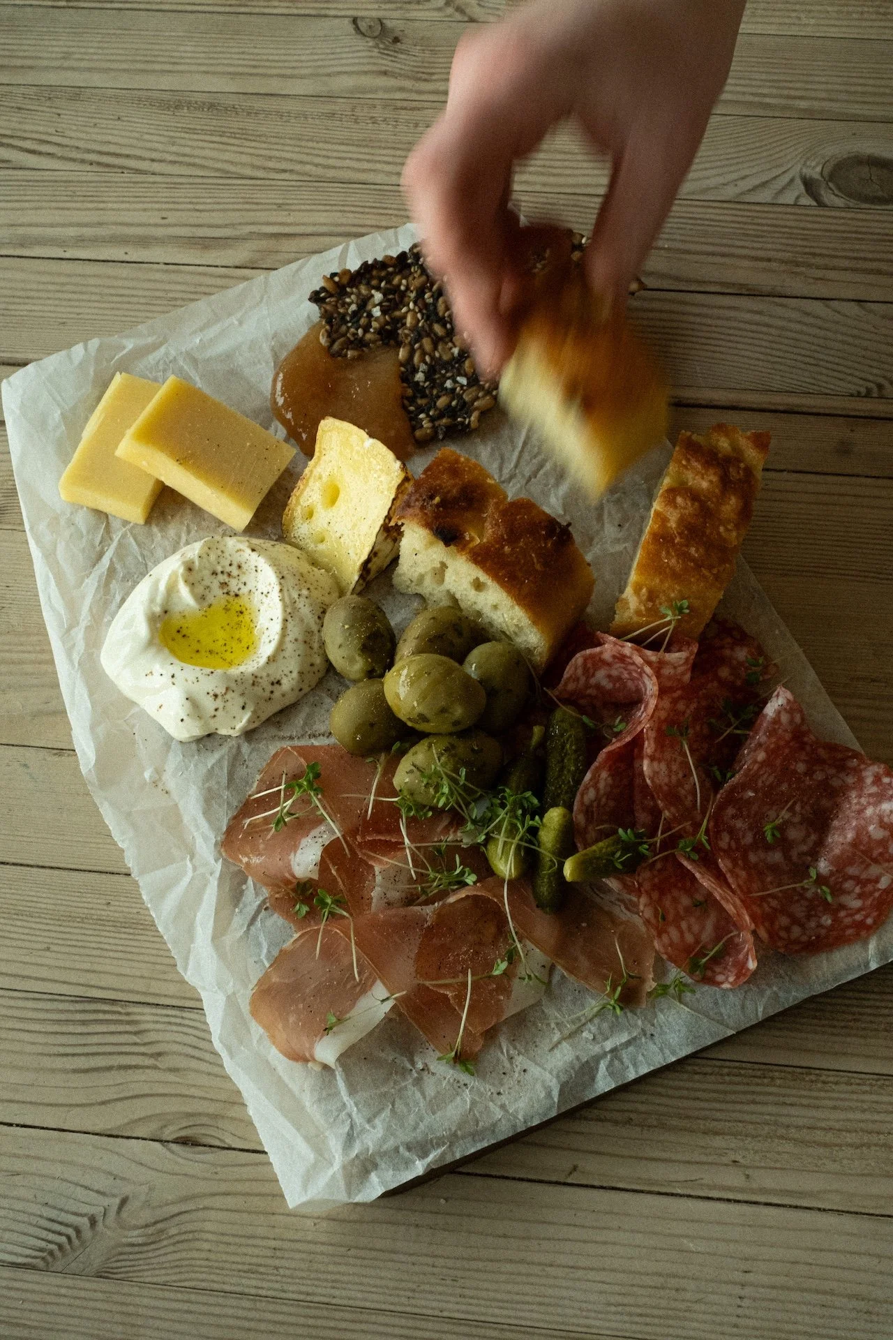 A charcuterie board with cheeses, cured meats, bread, green olives, pickles, and sliced cured meats garnished with microgreens, with a hand reaching for food.