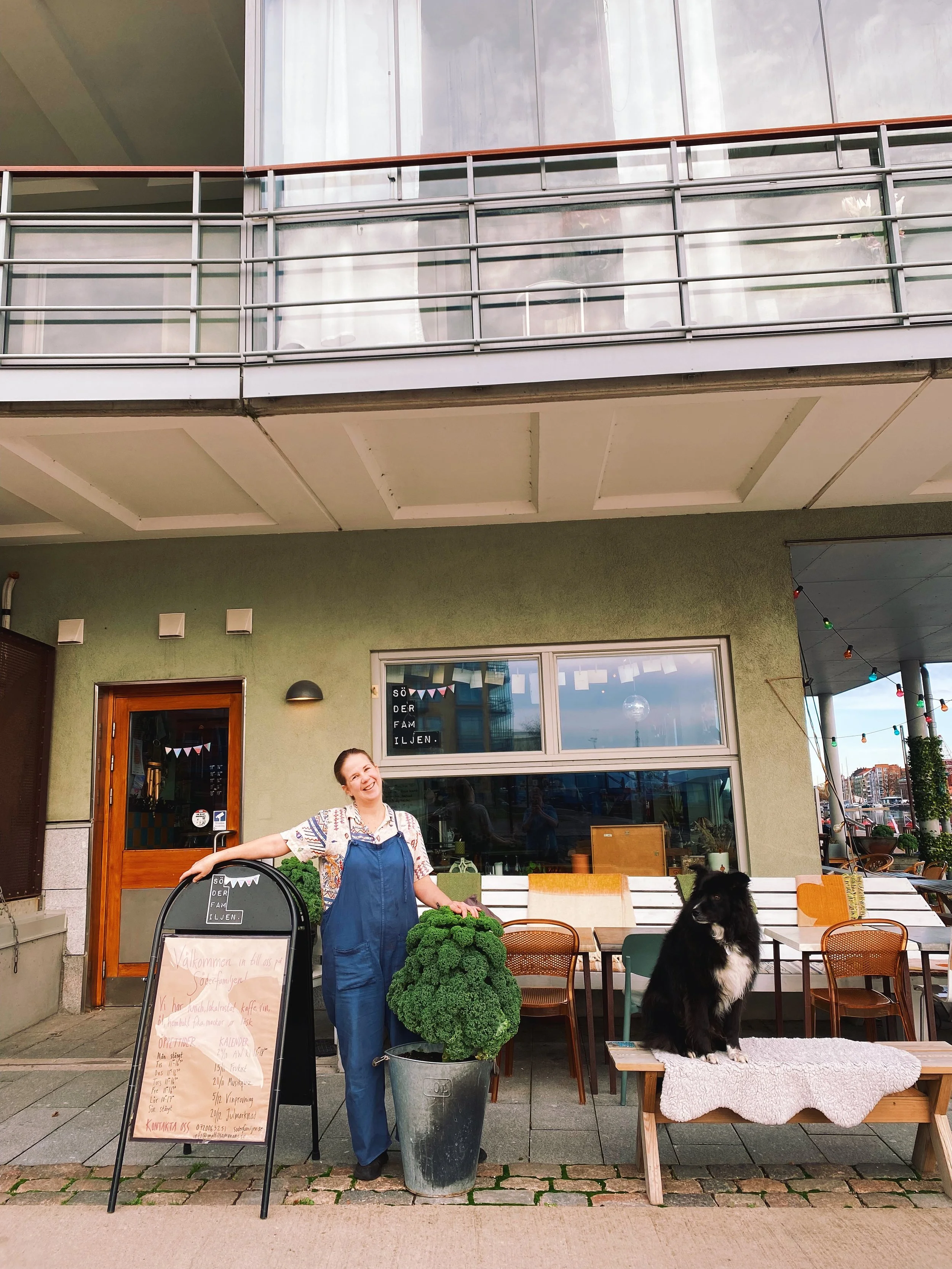Woman standing outside a cafe with a black and white dog on a small table in front. The woman is smiling and holding a large potted plant. There is a chalkboard menu and a large window behind them.