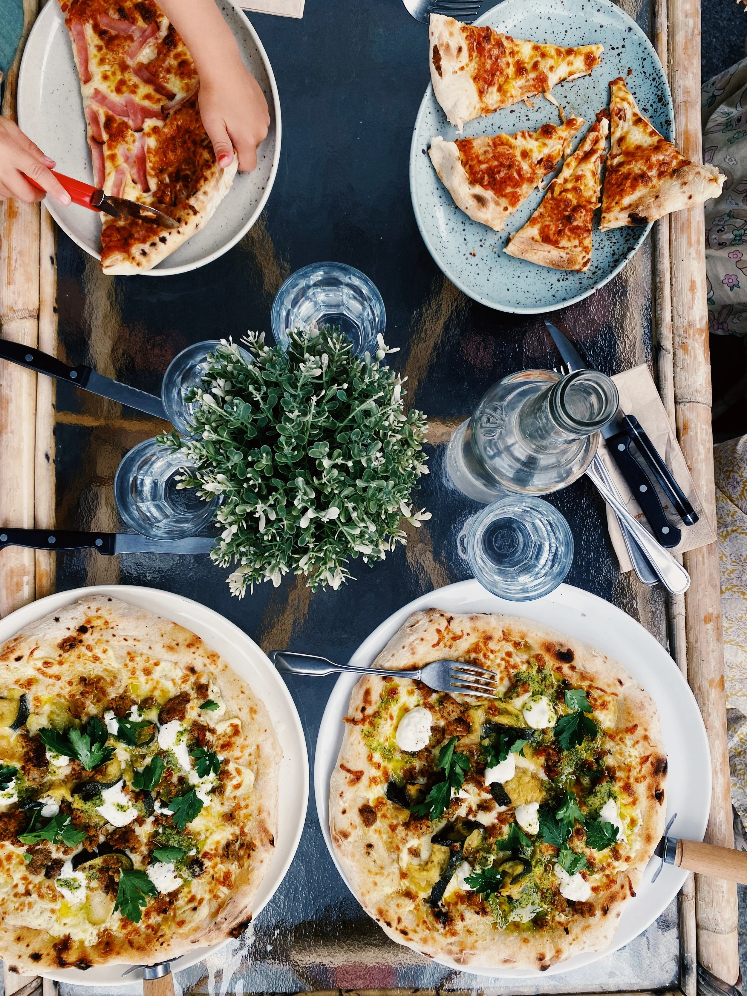 A dinner table with three pizza plates, some water glasses, a carafe, cutlery, and a small plant in the center.
