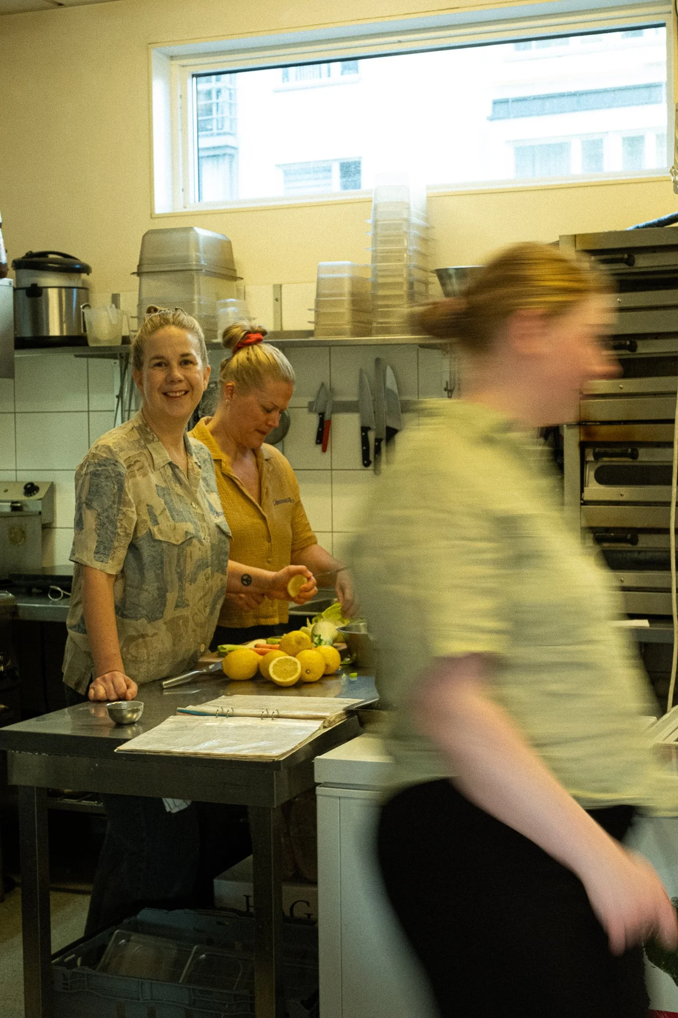 Three women working in a kitchen with lemons on the counter, two of them slicing lemons. One woman is smiling at the camera, while the others are focused on their work.