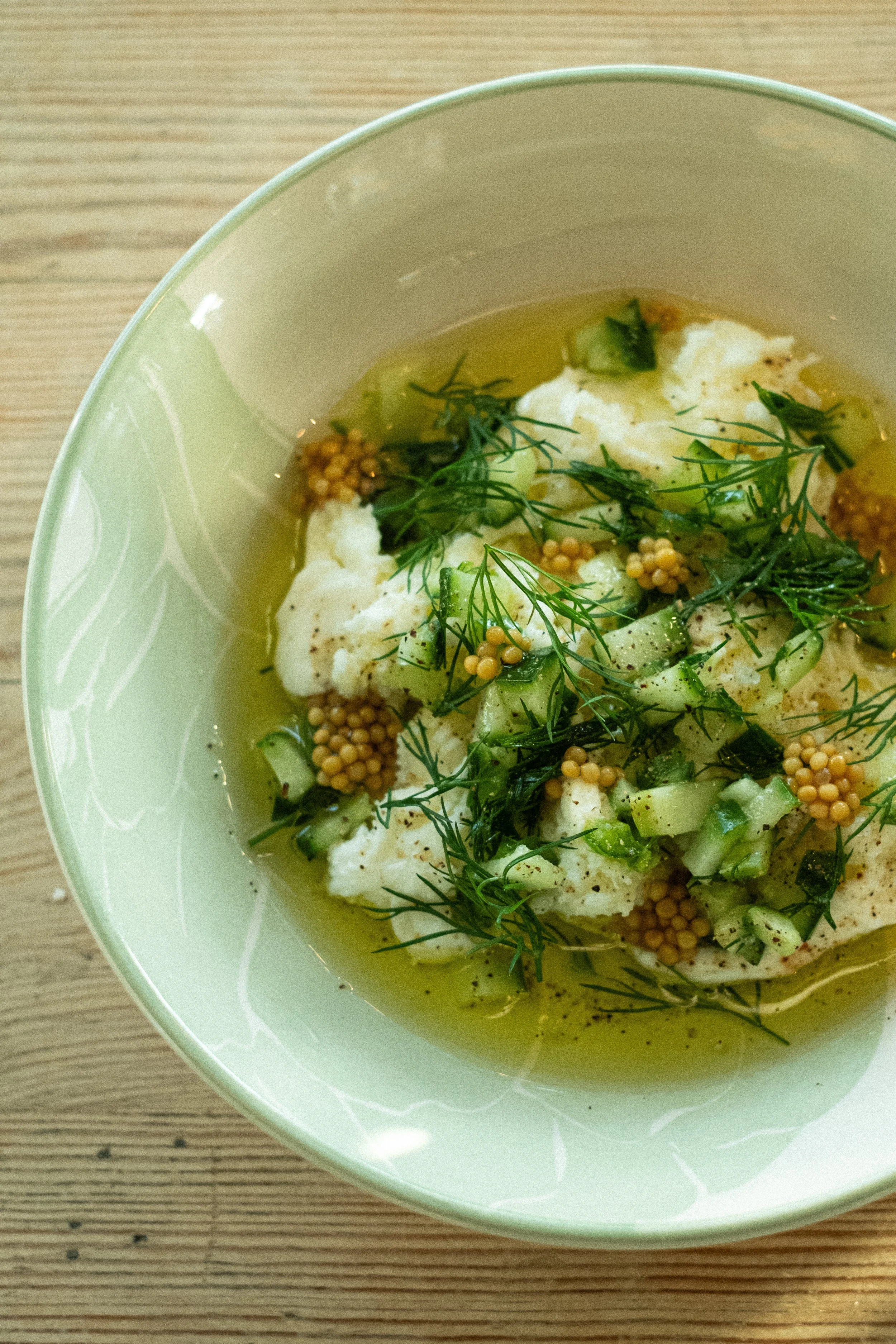 A bowl of cauliflower soup garnished with fresh herbs, mustard seeds, and chopped cucumbers.
