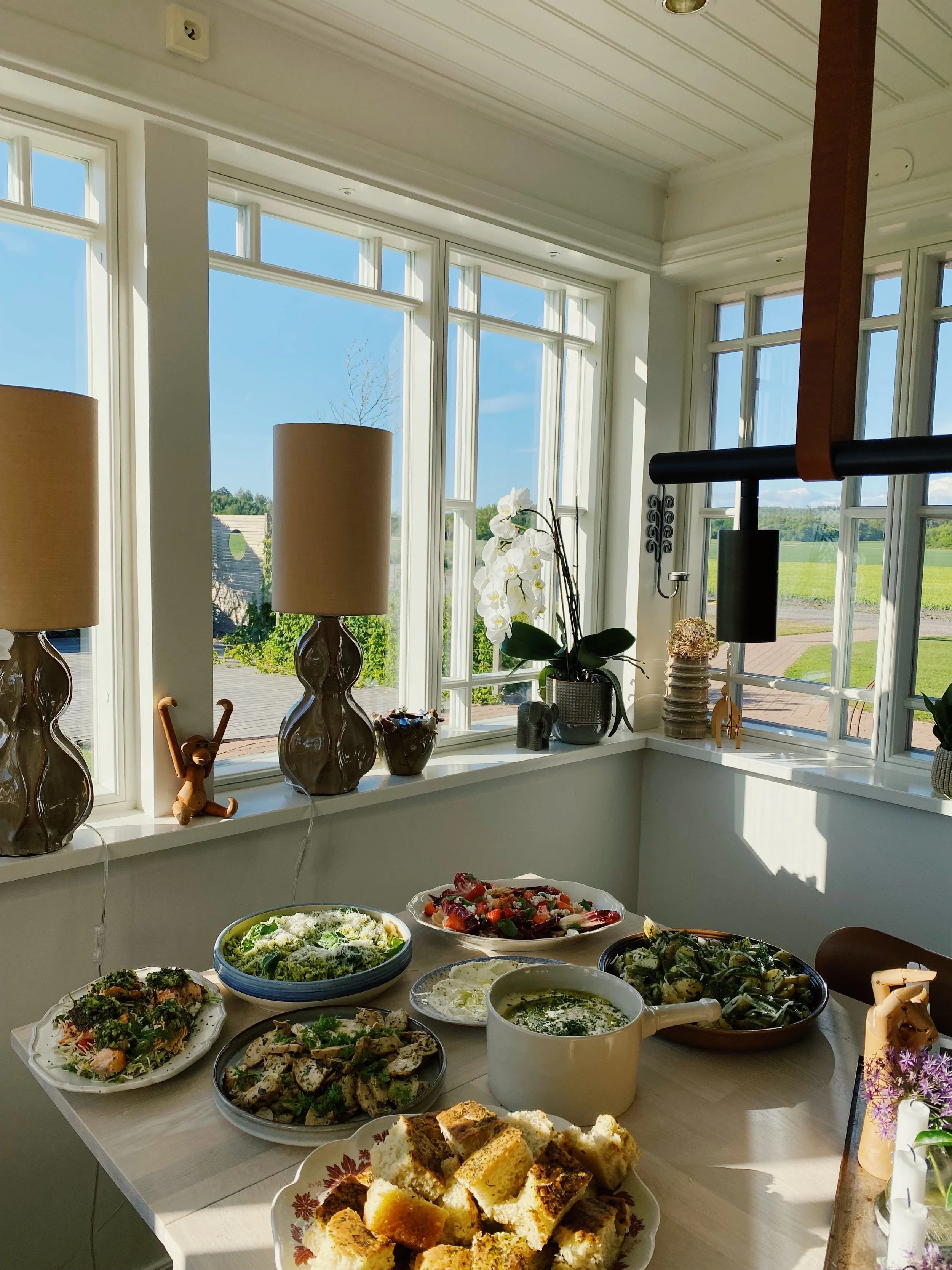 A table with various dishes of food, set in a bright room with large windows showing a sunny outdoor view. The table has bread, salads, and other dishes, with sunlight streaming in.