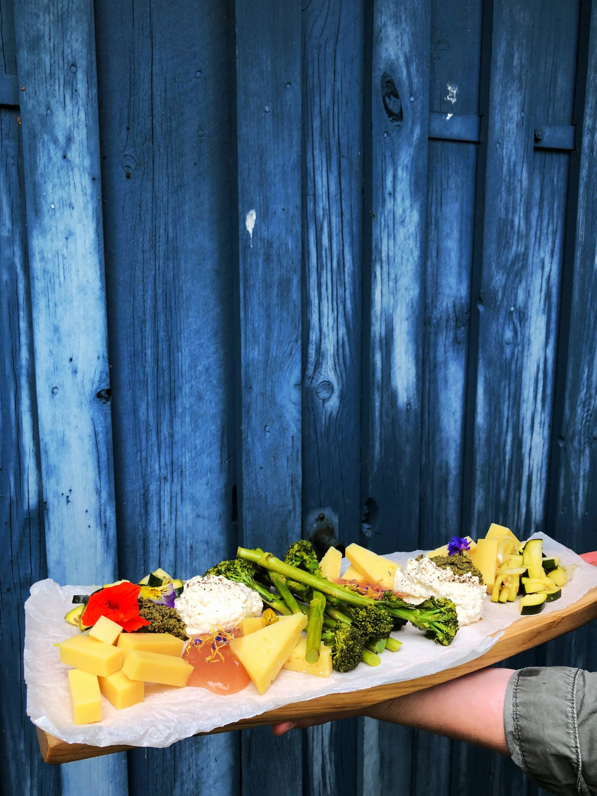 A hand holding a wooden platter with assorted cheeses, broccoli, garnishes, and edible flowers against a blue wooden fence.