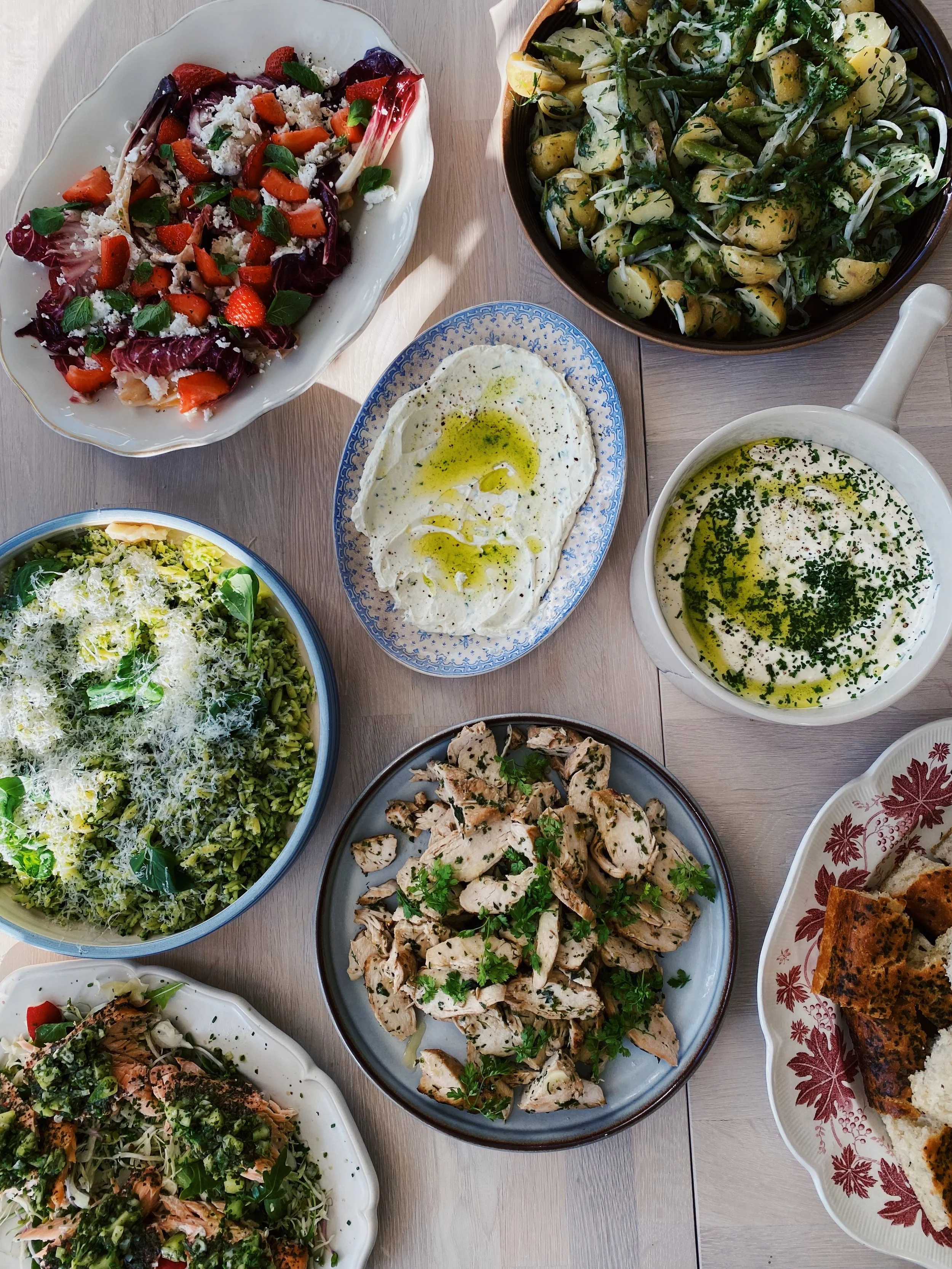 Assorted dishes including a strawberry and vegetable salad, a potato salad with herbs, a creamy garlic dip drizzled with olive oil, a green vegetable pasta salad, a broccoli and cheese salad, grilled chicken slices garnished with herbs, a spinach and salmon salad, and a dish of grilled flatbread.