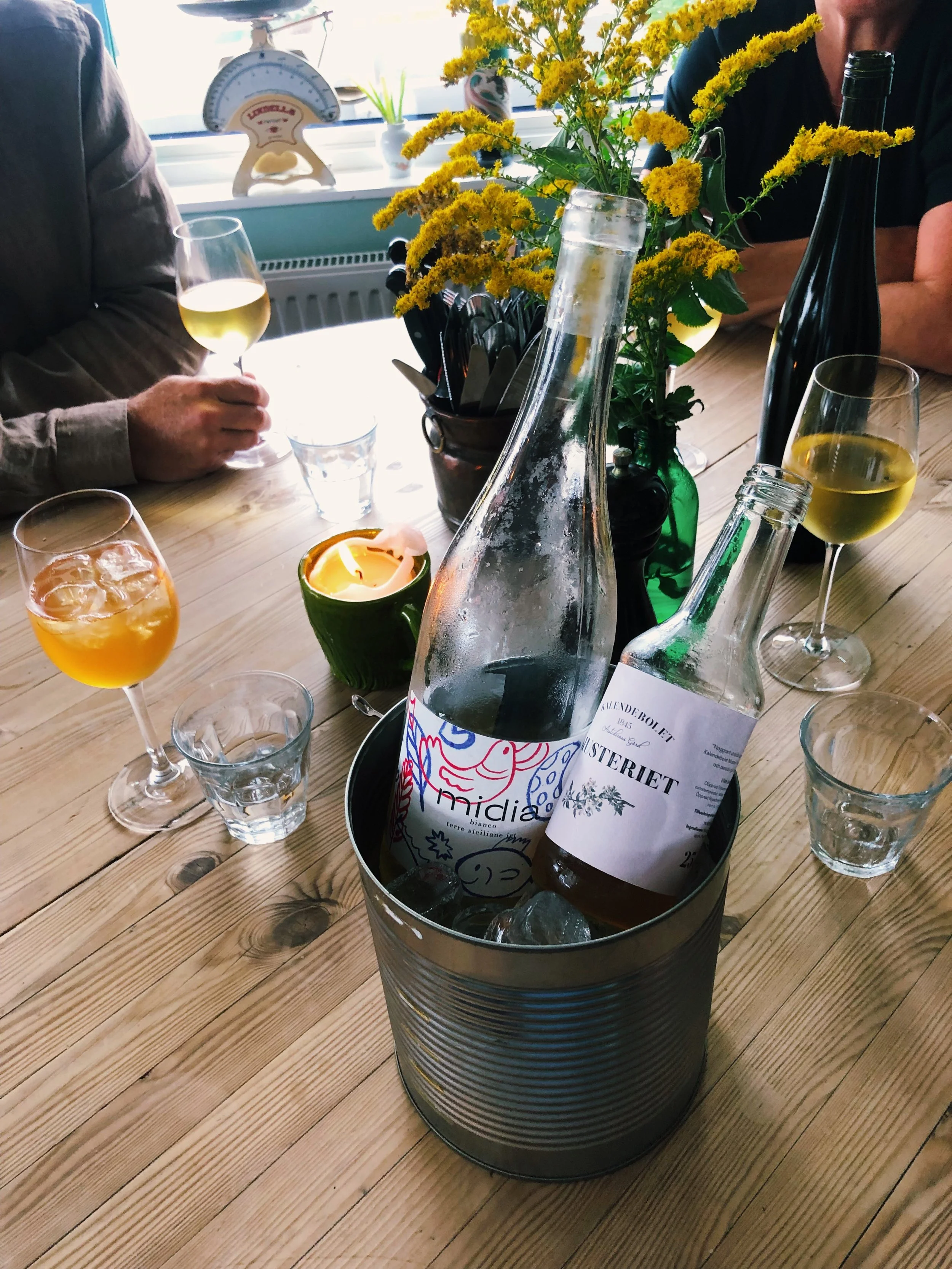 Table with a metal bucket containing two glass bottles of beverages, one labeled "mindalia" and the other "Osteriet", surrounded by glasses of white wine, a small green candle holder, and a flower arrangement with yellow flowers. In the background, there are people, a window, a scale, and various utensils.