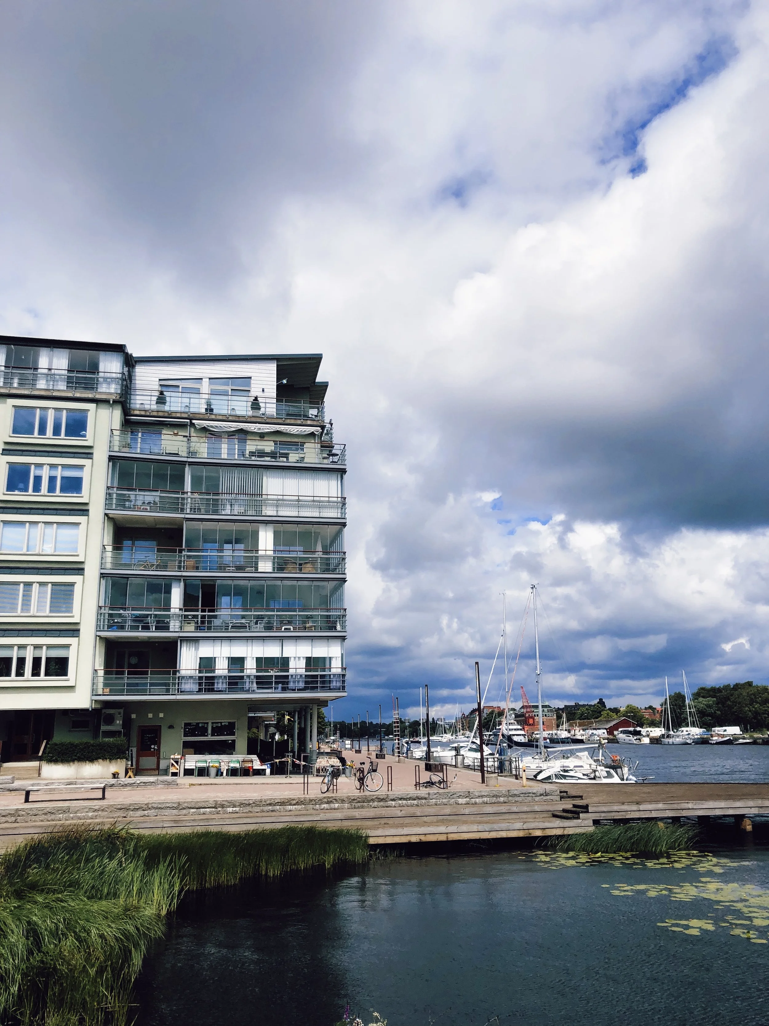 Modern apartment building near a marina with boats docked, some bicycles parked outside, and a cloudy sky overhead.