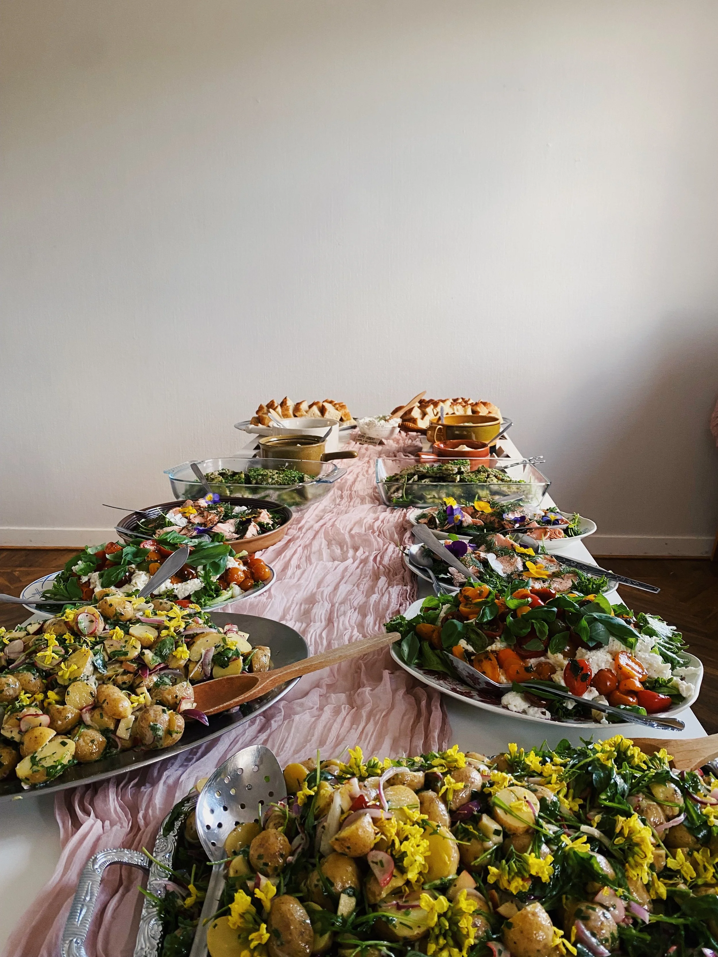 A table with various salads and desserts, including potato salad, greens, and pies, decorated with edible flowers and a pink tablecloth.
