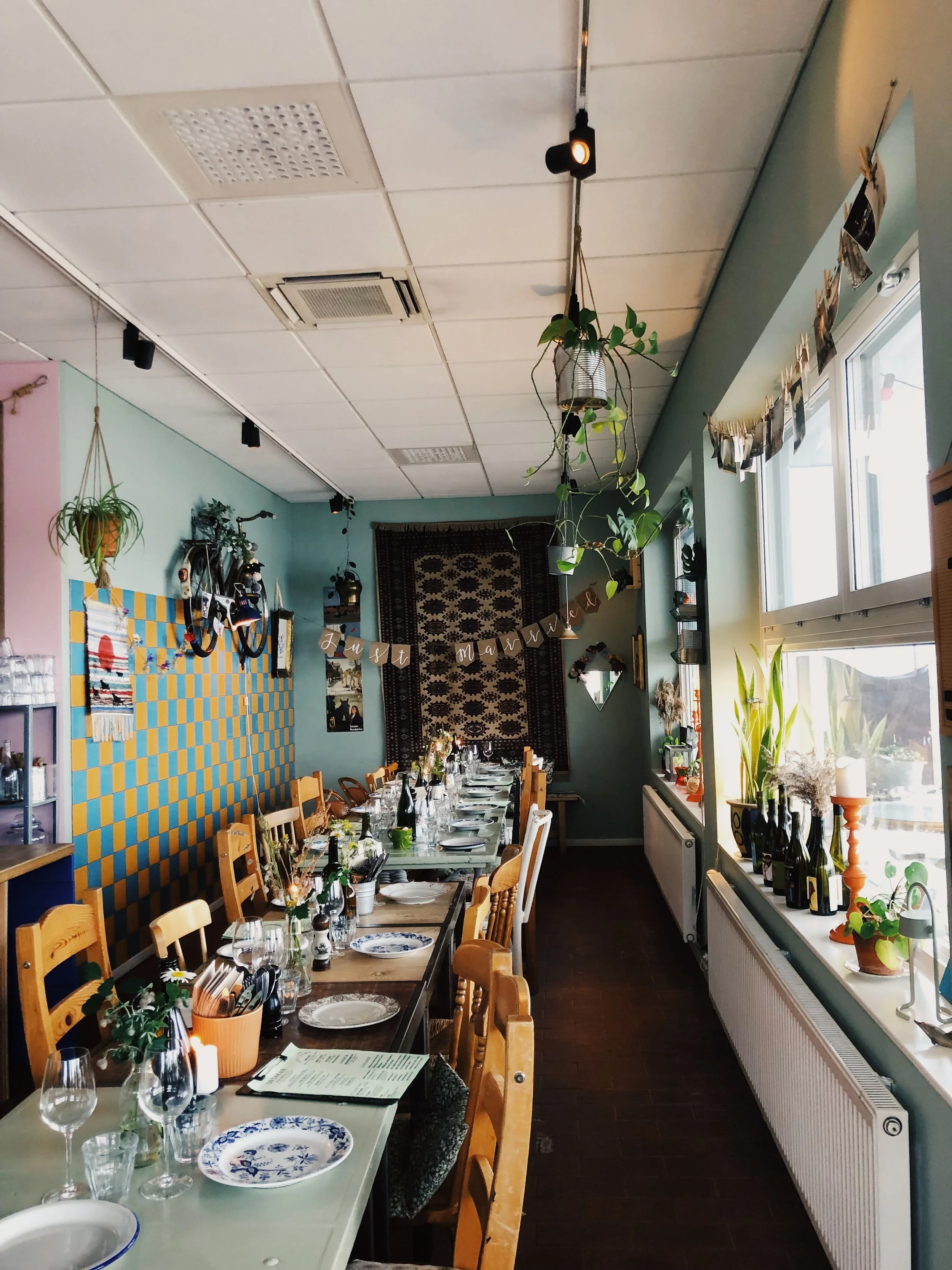Interior of a decorated restaurant with a long dining table set with plates, glasses, and cutlery. There are potted plants on the windowsill and hanging from the ceiling. A banner reading 'Just Married' is displayed on the back wall.