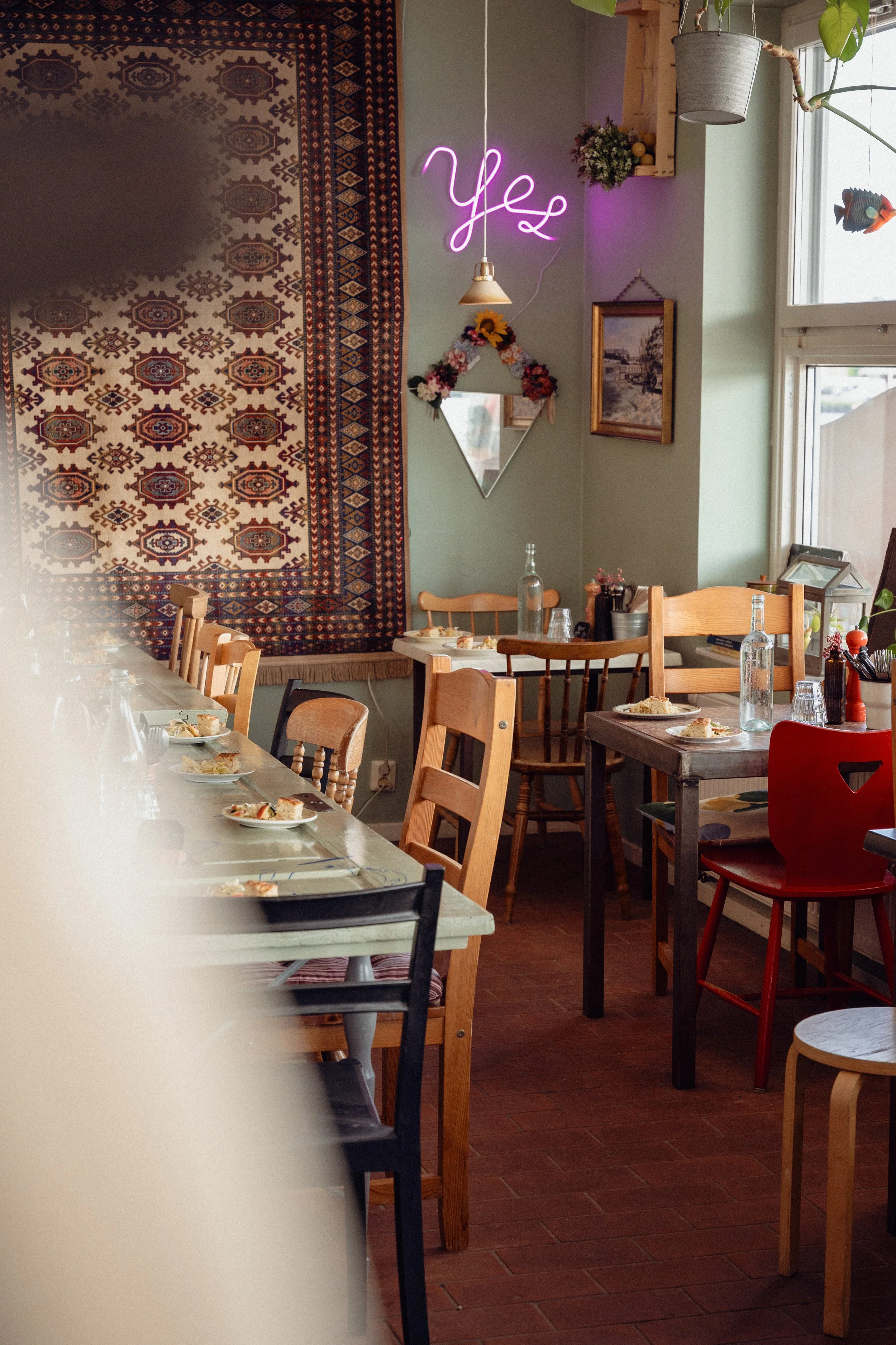 Inside a cozy restaurant with wooden chairs, some around a long table with place settings, and colorful decor including a neon 'Yes' sign on the wall, a traditional patterned rug, framed pictures, and hanging plants near windows.