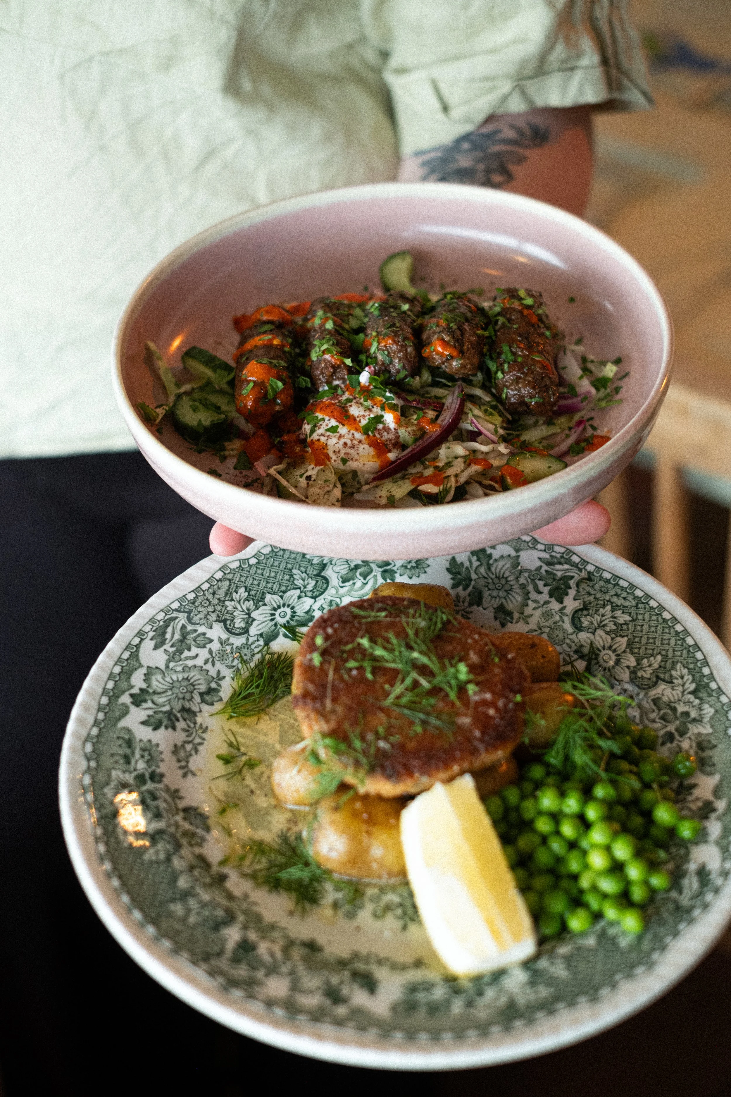 Two plates of food: the top bowl contains kebabs served over a salad with cucumber, red onion, and herbs, garnished with sauce and seasoning. The bottom plate features a cooked meat patty with sauce, accompanied by green peas, a lemon wedge, and herbs.