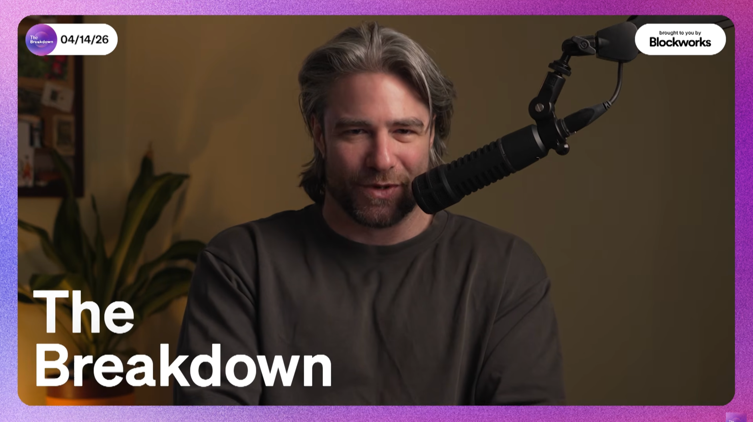 A man with gray hair and beard, wearing a dark shirt, speaking into a microphone during a podcast titled 'The Breakdown,' with a plant in the background.