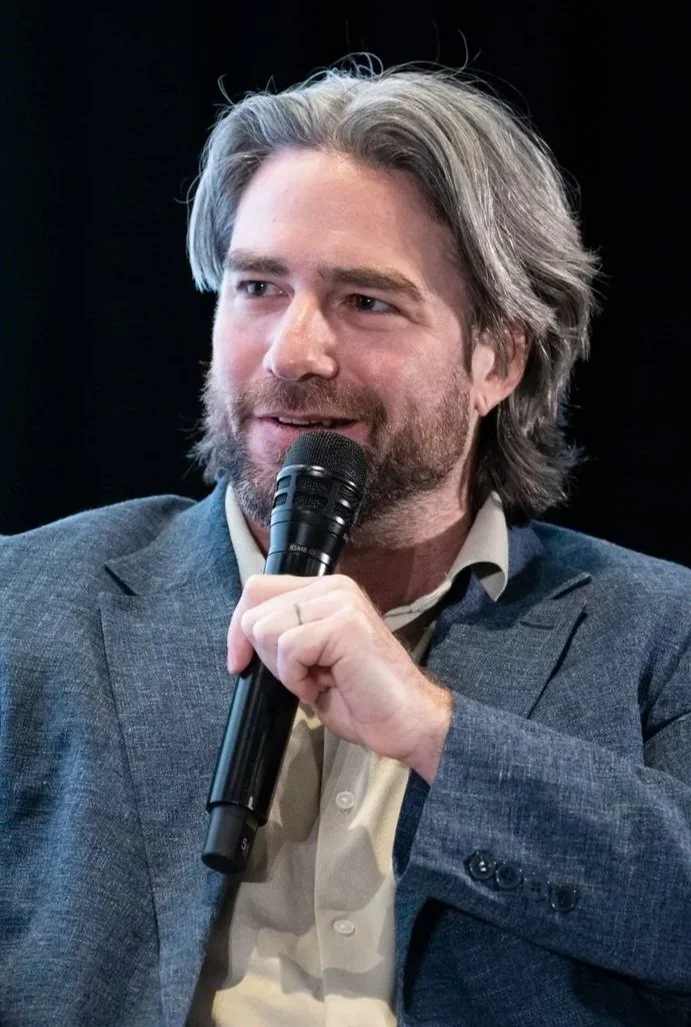 A man with shoulder-length gray and brown hair and a beard, sitting and speaking into a microphone, wearing a gray blazer and a light-colored shirt with a black background.