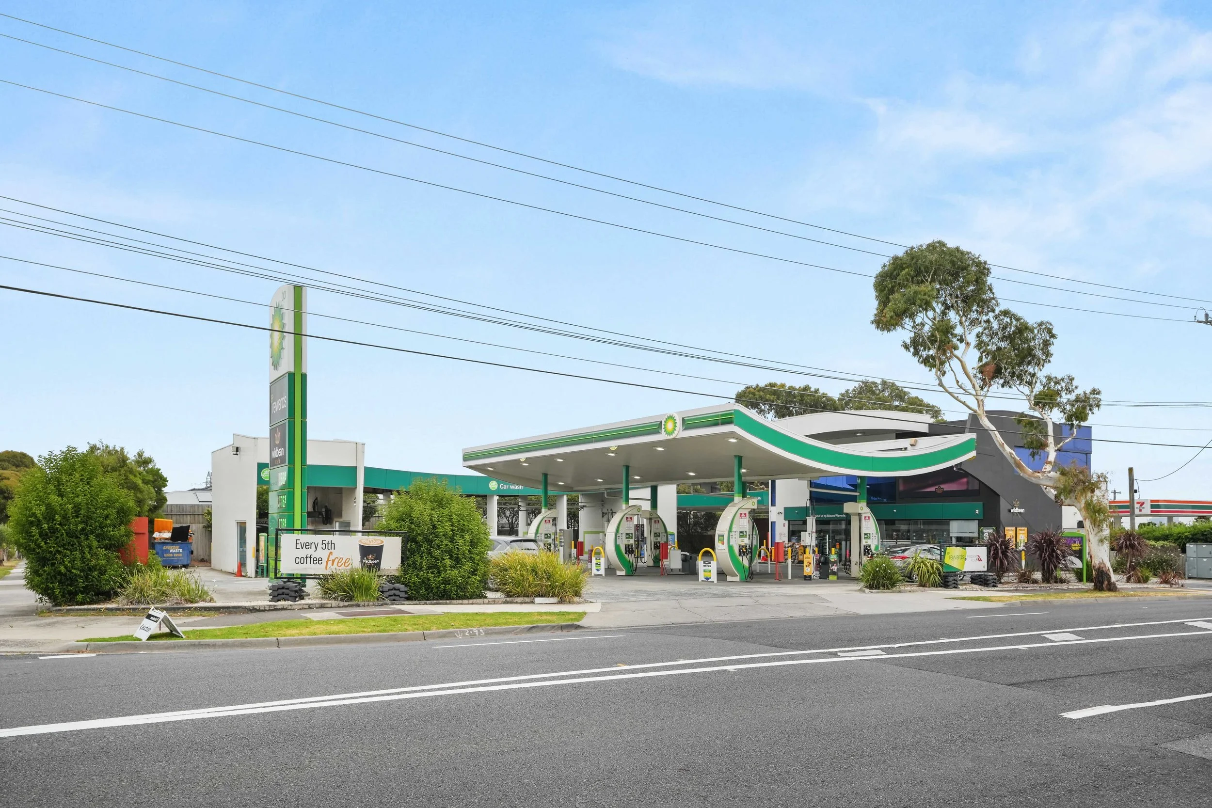 A BP gas station with multiple fueling pumps under a canopy, surrounded by greenery, with a sign advertising every 5th coffee free and a nearby 7-Eleven store in the background.