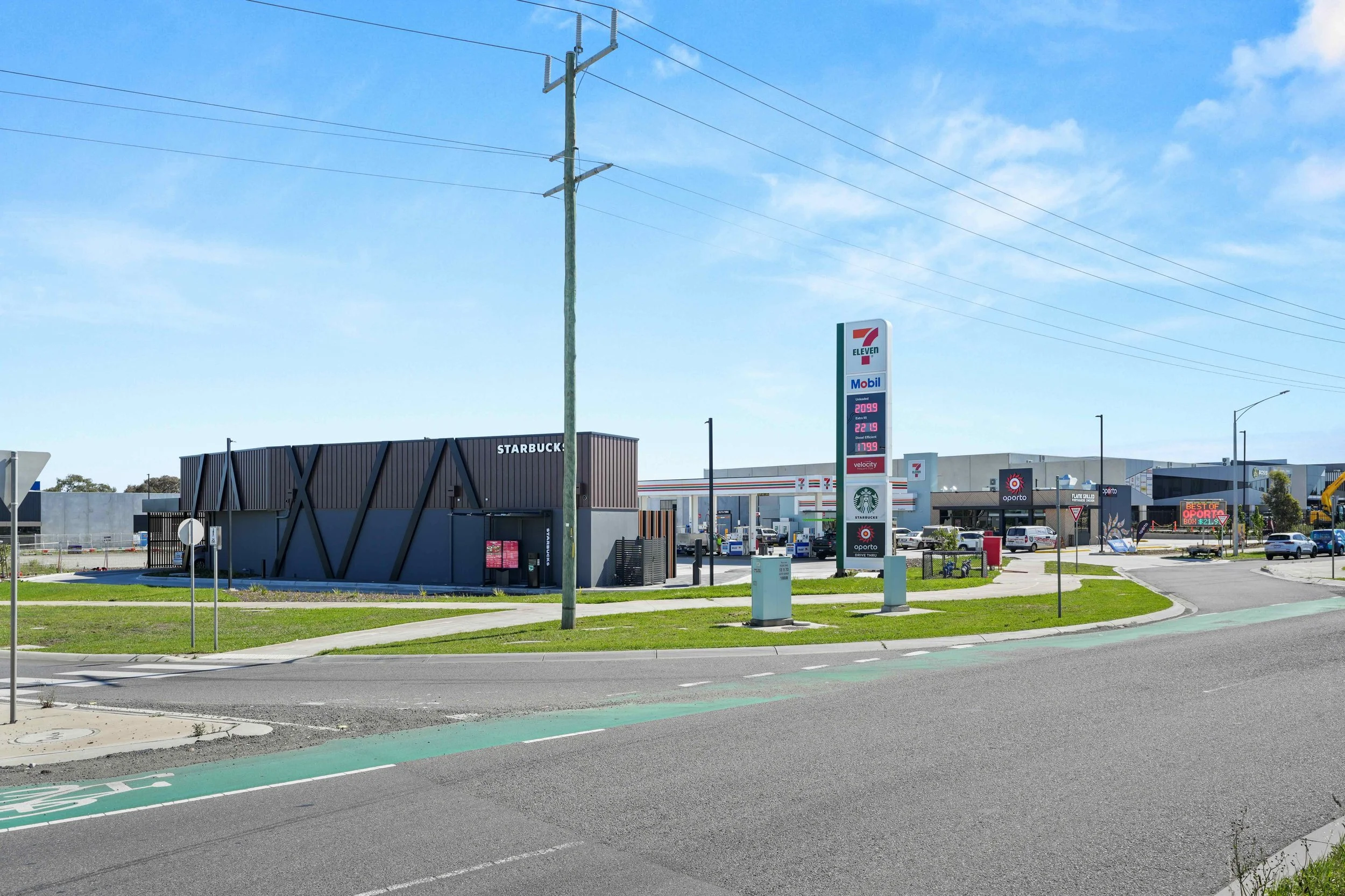 A gas station with a 7-Eleven sign, convenience store, and a Starbucks located nearby. The gas prices are displayed on a tall signboard, and there are parked cars and a few bicycles around. The sky is blue with some clouds, and the area is well-maint