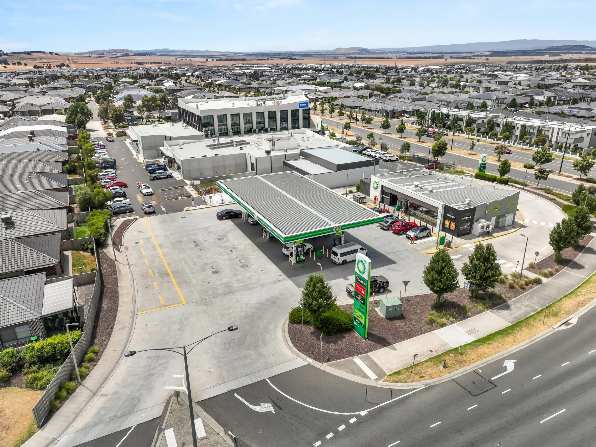 A gas station with a convenience store, situated near residential houses in a suburban area. Multiple cars are parked at the gas pumps and in the parking lot, with a large sign displaying the BP logo and various fuel prices. The background features a