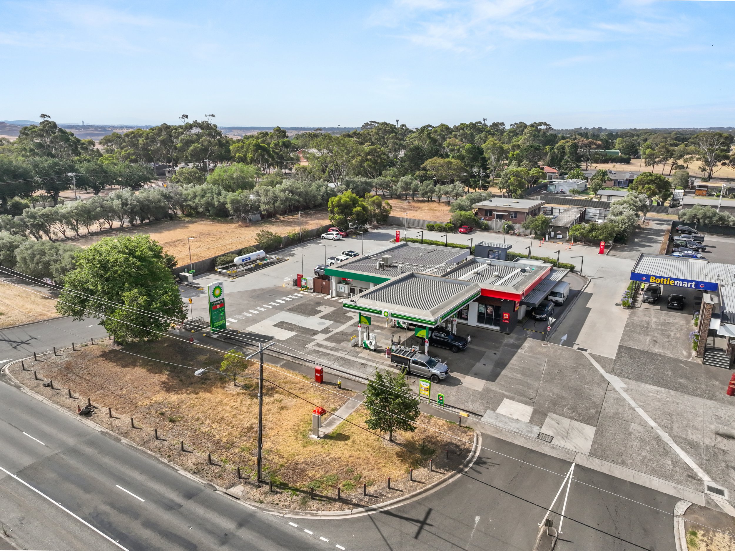 An aerial view of a BP gas station with a convenience store, multiple fuel pumps, parked cars, and surrounding trees and open land in the background.