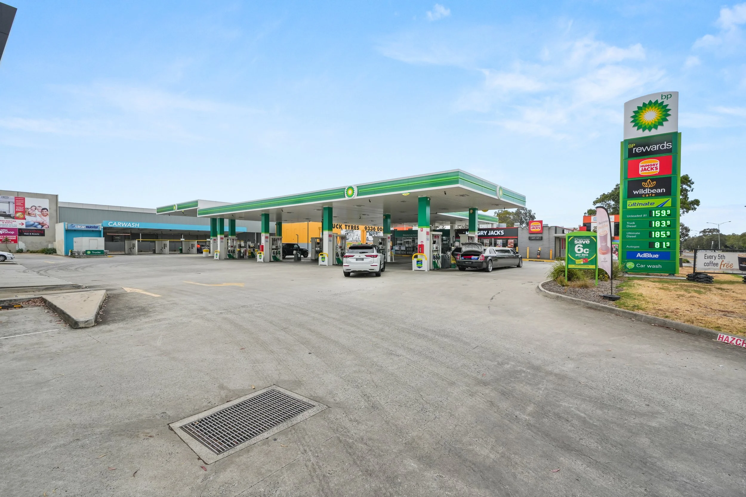 A BP gas station with multiple fuel pumps, cars refueling, and a tall sign displaying fuel prices and nearby stores, under a clear blue sky.