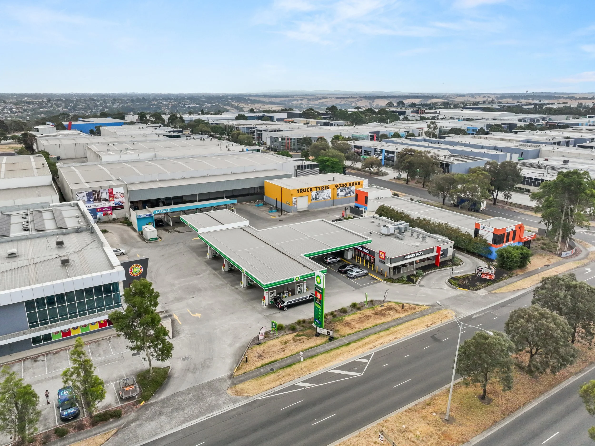 Aerial view of a shopping center with a BP petrol station, Hungry Jack's, and other retail stores, surrounded by trees and roads in a commercial area.