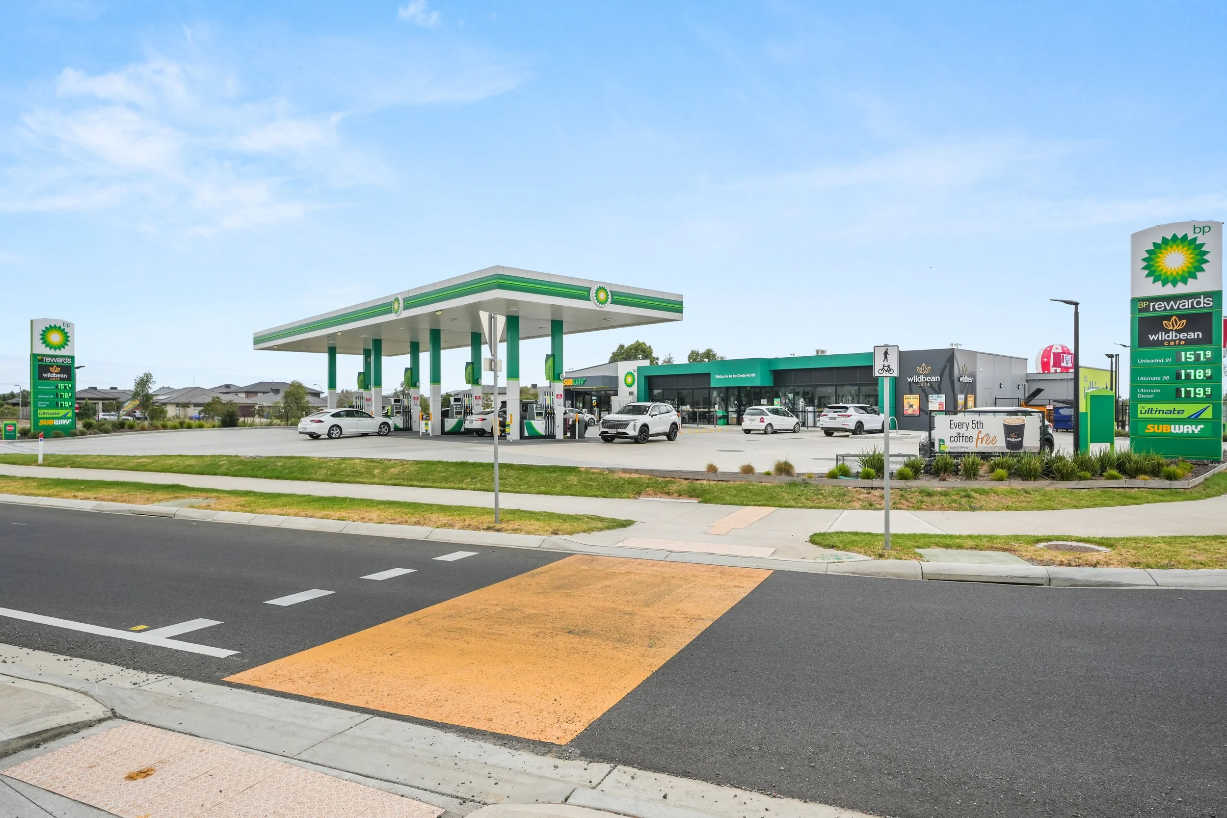 Gas station with green and white canopy, multiple fuel pumps, cars parked, and a convenience store in the background. Price signboard displaying fuel prices, sidewalk, and street in the foreground.