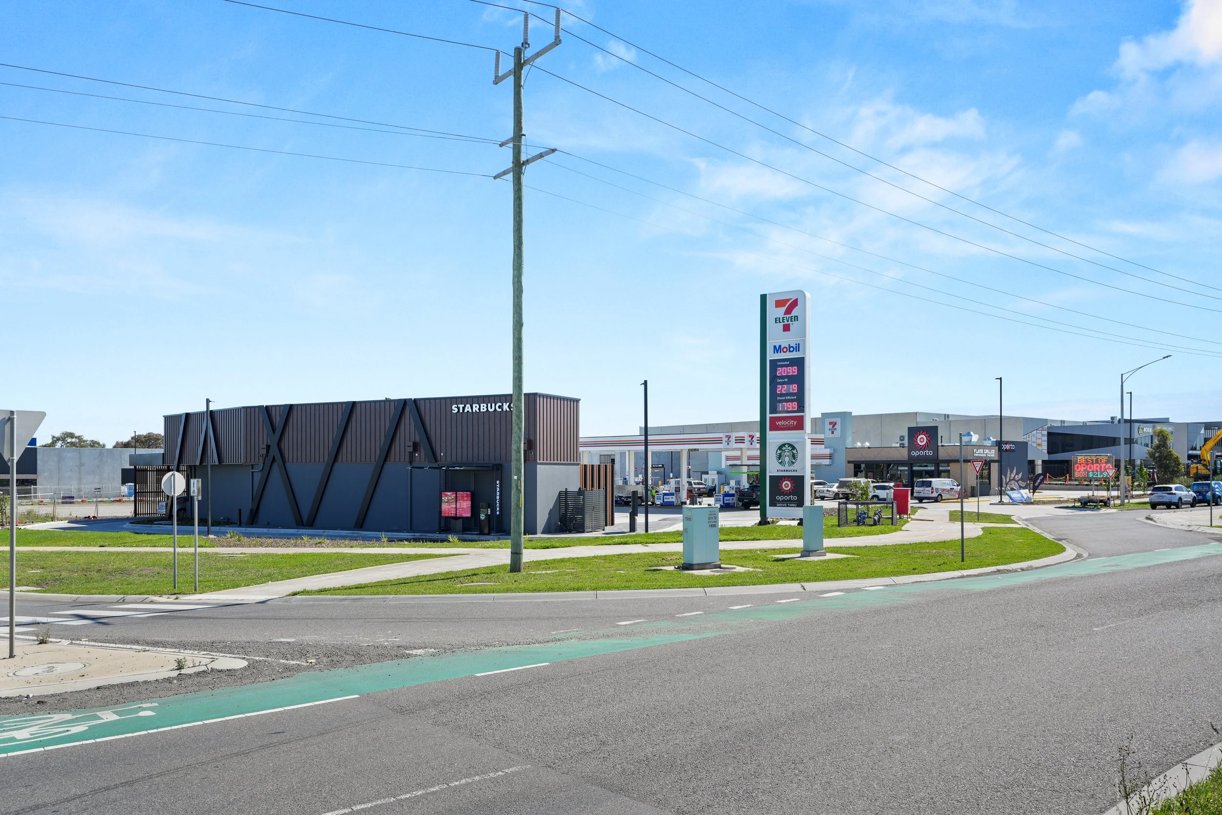 A roadside scene featuring a 7-Eleven convenience store and a Starbucks building, with fuel prices displayed on a sign, a petrol station with multiple bays, and vehicles parked around, under a clear blue sky.