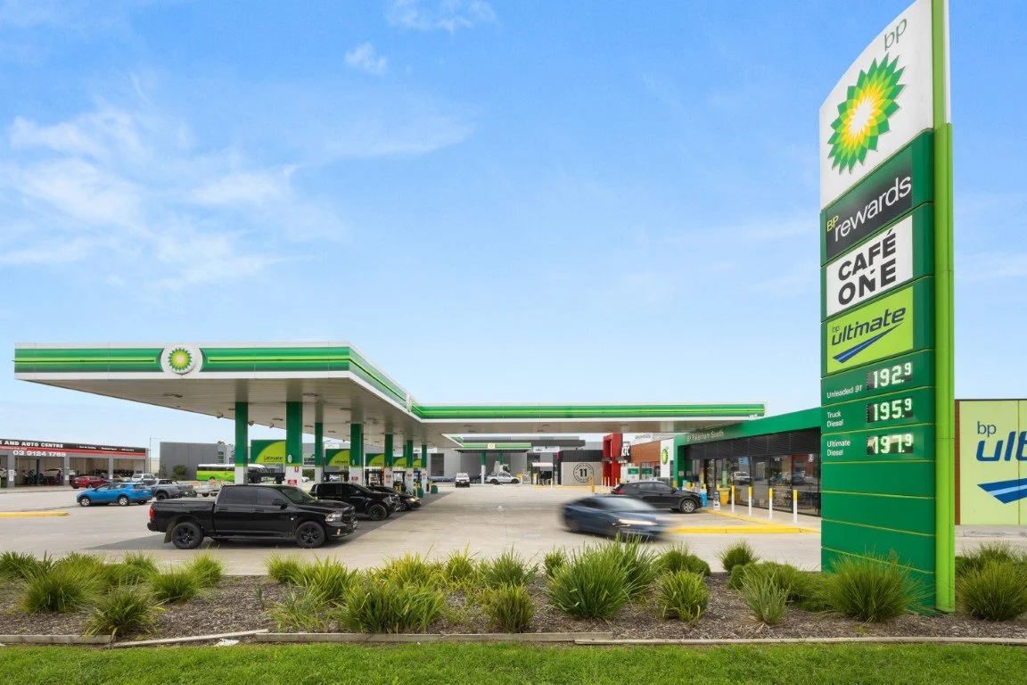 Gas station with BP branding, fuel prices listed on a sign, several cars parked and refueling, a convenience store on the side, and a clear blue sky.