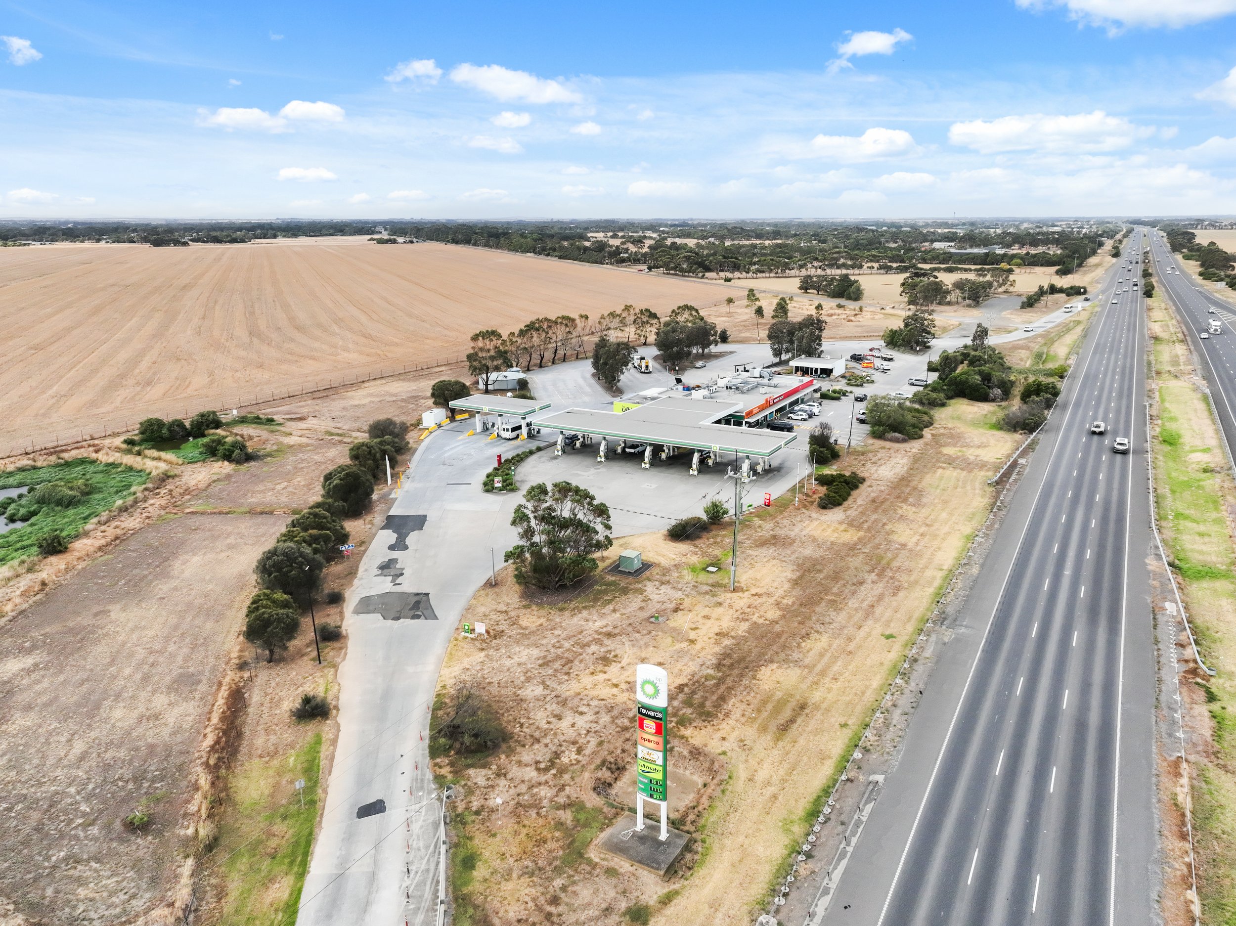 An aerial view of a BP gas station and convenience store situated beside a highway with multiple lanes. The gas station has a large canopy over fueling pumps, with parked cars and trucks nearby. The surrounding area includes dry fields and patches of greenery, with a long straight highway extending into the distance under a partly cloudy sky.