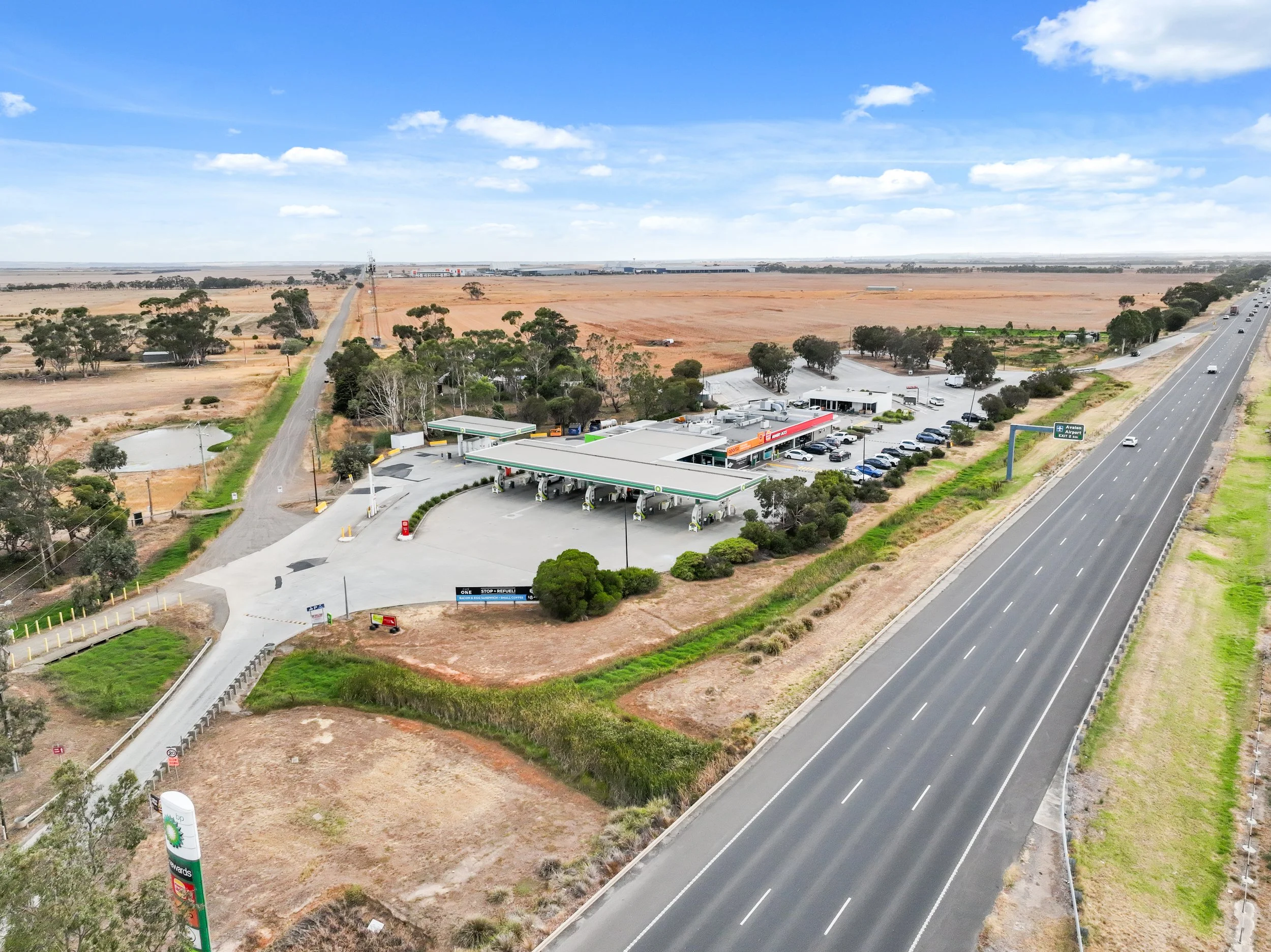 A BP gas station with multiple fueling pumps, a convenience store, and a parking lot, located next to a multi-lane highway in a semi-rural area, with open farmland and sparse trees in the background.