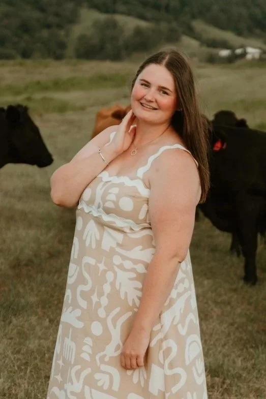 A smiling woman with long brown hair wearing a beige and white patterned dress standing outdoors in a grassy field with cows in the background.