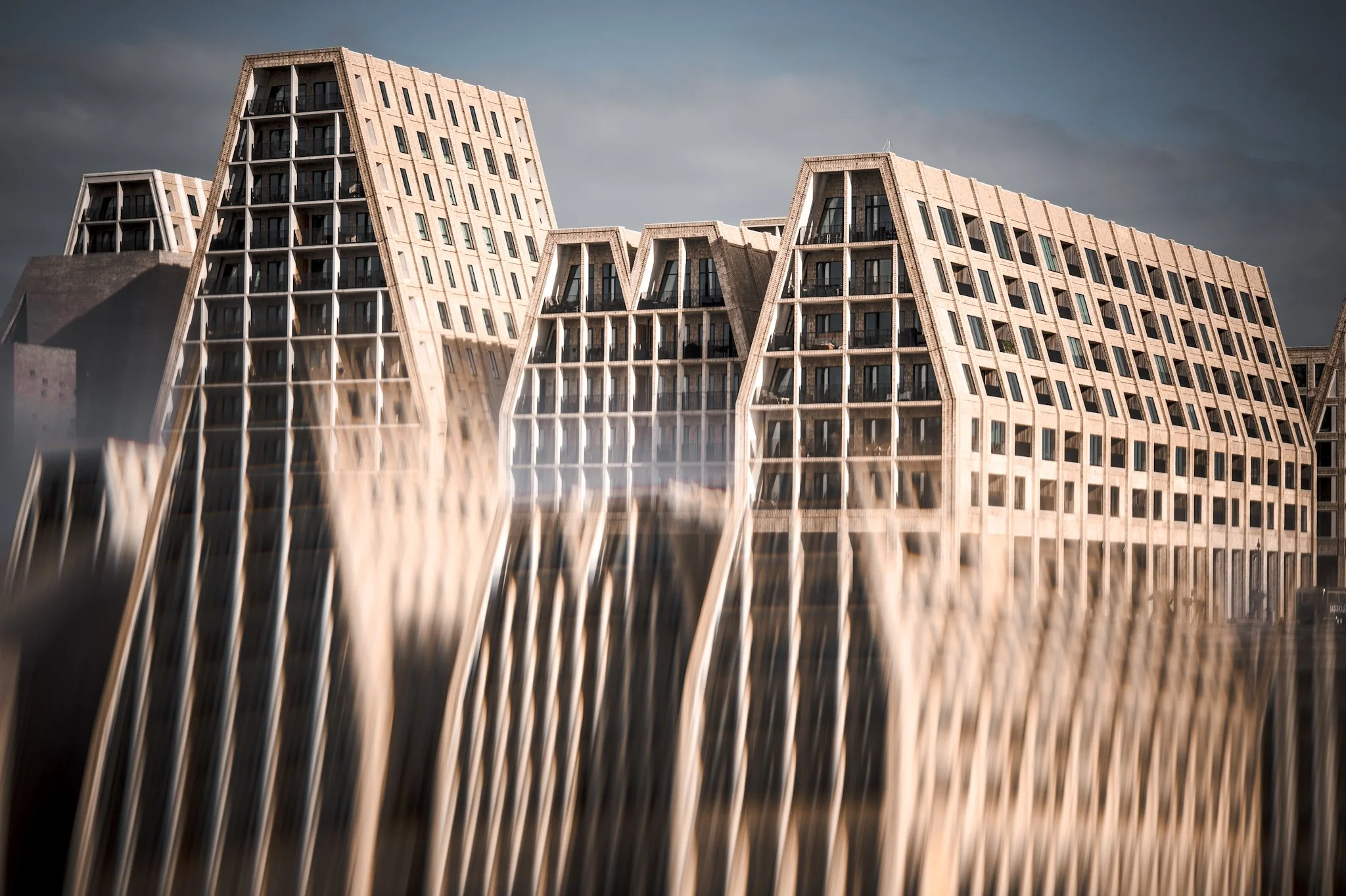Modern multi-story buildings with angular facades reflect in water, under a cloudy sky.