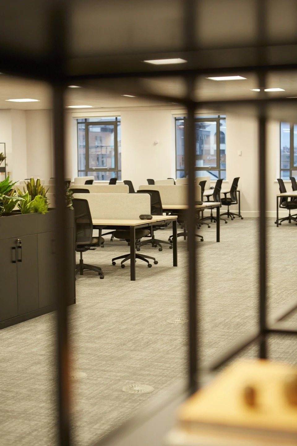 Empty office with rows of desks, chairs, and large windows overlooking city buildings, partly viewed through a black metal grid or partition.