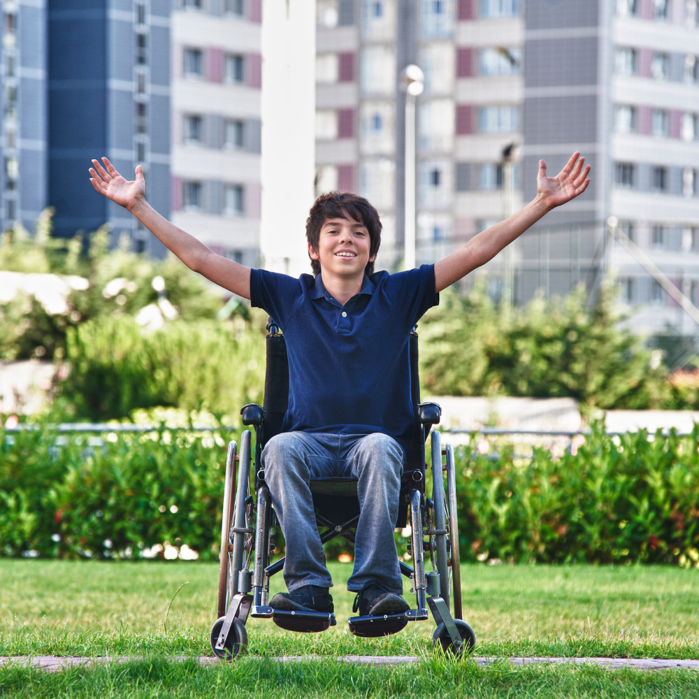 A young boy sitting in a wheelchair outdoors with arms raised, smiling, with tall buildings and greenery in the background.