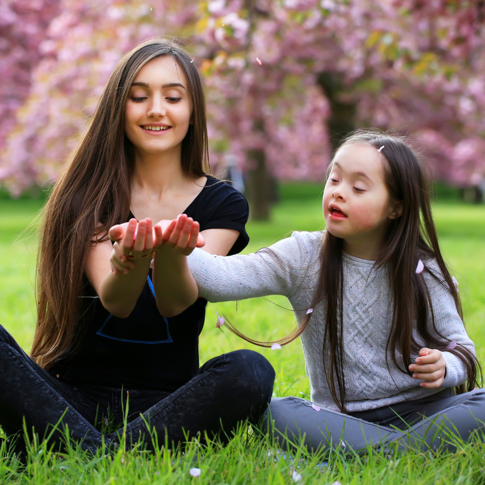 A young woman and a little girl sitting on grass under pink cherry blossom trees, exchanging flower petals in a park.