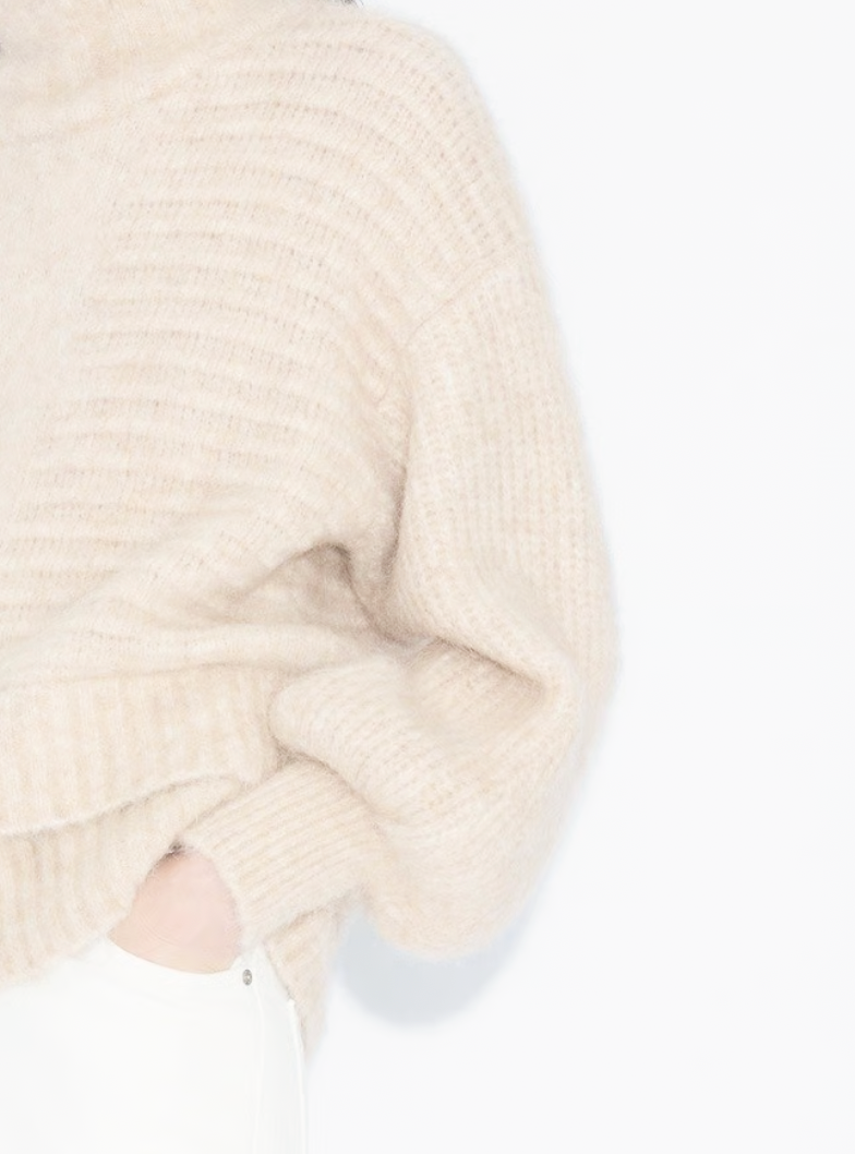 Close-up of a person wearing a cream-colored, chunky knit sweater with a pocket, standing against a white background.