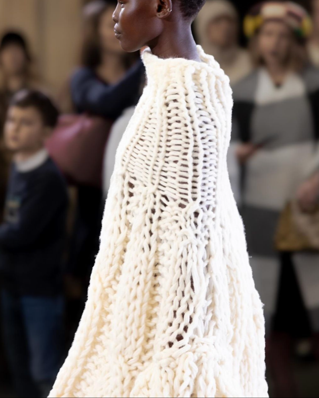 Model walking on runway in chunky, oversized cream knit sweater during fashion show, with blurred audience watching.