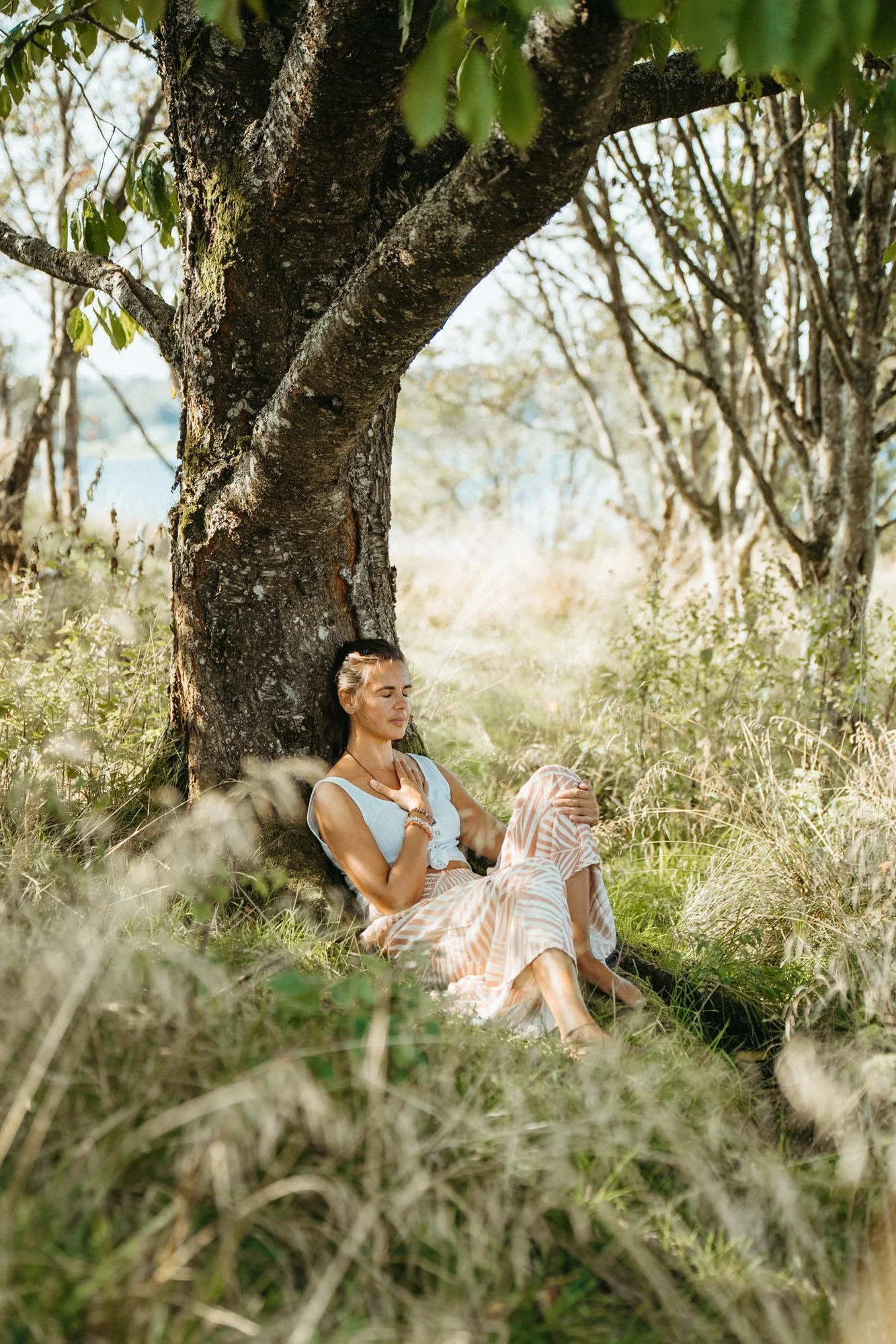 A woman sitting under a large tree in a grassy field, eyes closed, appearing to relax or meditate.