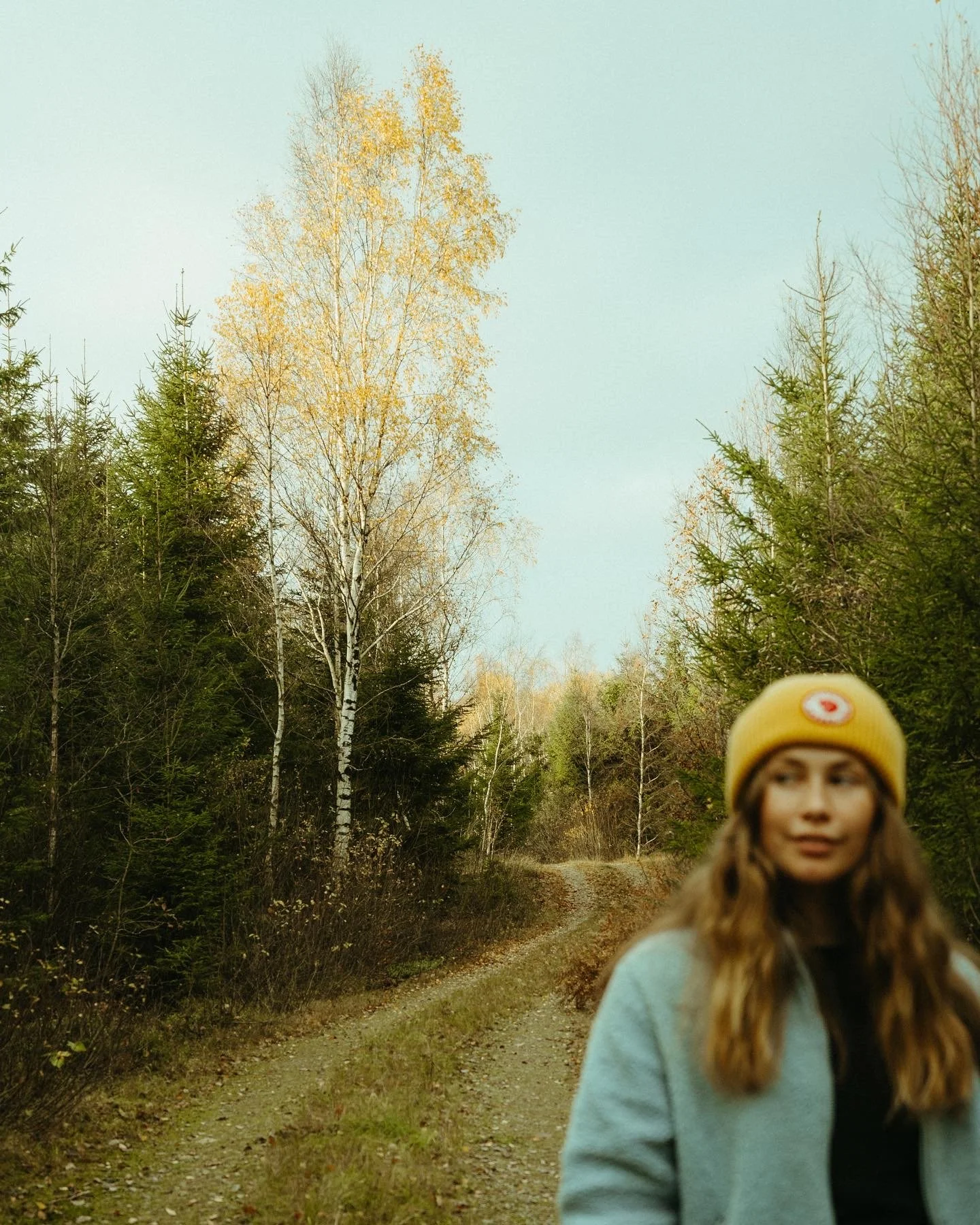 A woman with long wavy hair wearing a yellow beanie and a light gray coat standing on a dirt trail in a forest during fall.