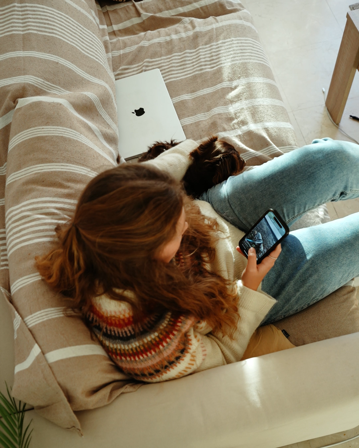 A woman with long, curly hair sitting on a beige striped couch, holding a smartphone, with a small dog next to her and a closed Apple laptop on the couch.